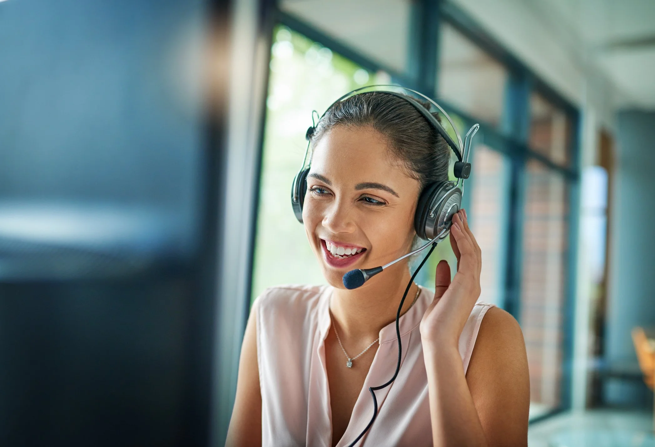 A young woman smiling while wearing a headset with a microphone, sitting in front of a computer in a bright, modern office or workspace with large windows.