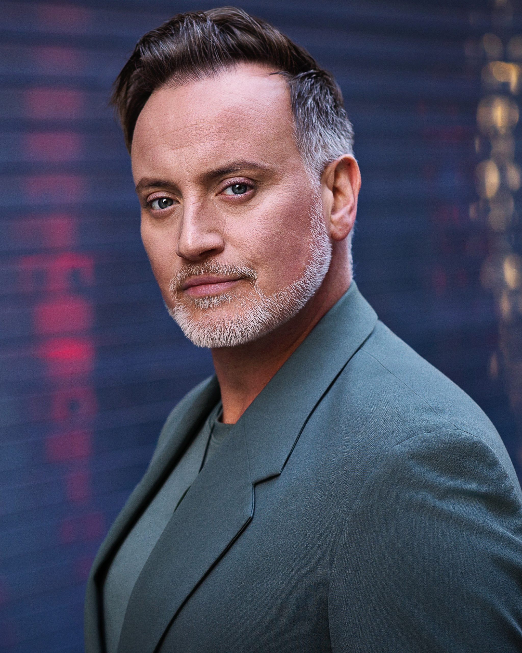 Close-up portrait of Hayden Tee with short, dark hair and a gray beard, wearing a green jacket and gray shirt, standing in front of a dark background with blurred lights.