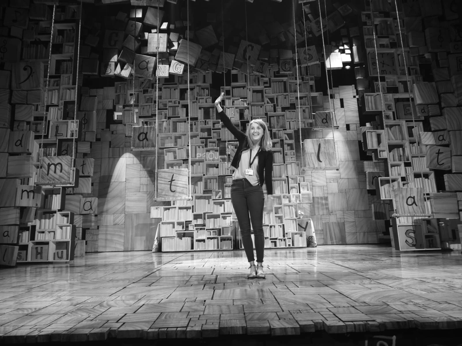 Liz Flint smiling and waving on the Matilda West End stage in front of a backdrop floating books and letters