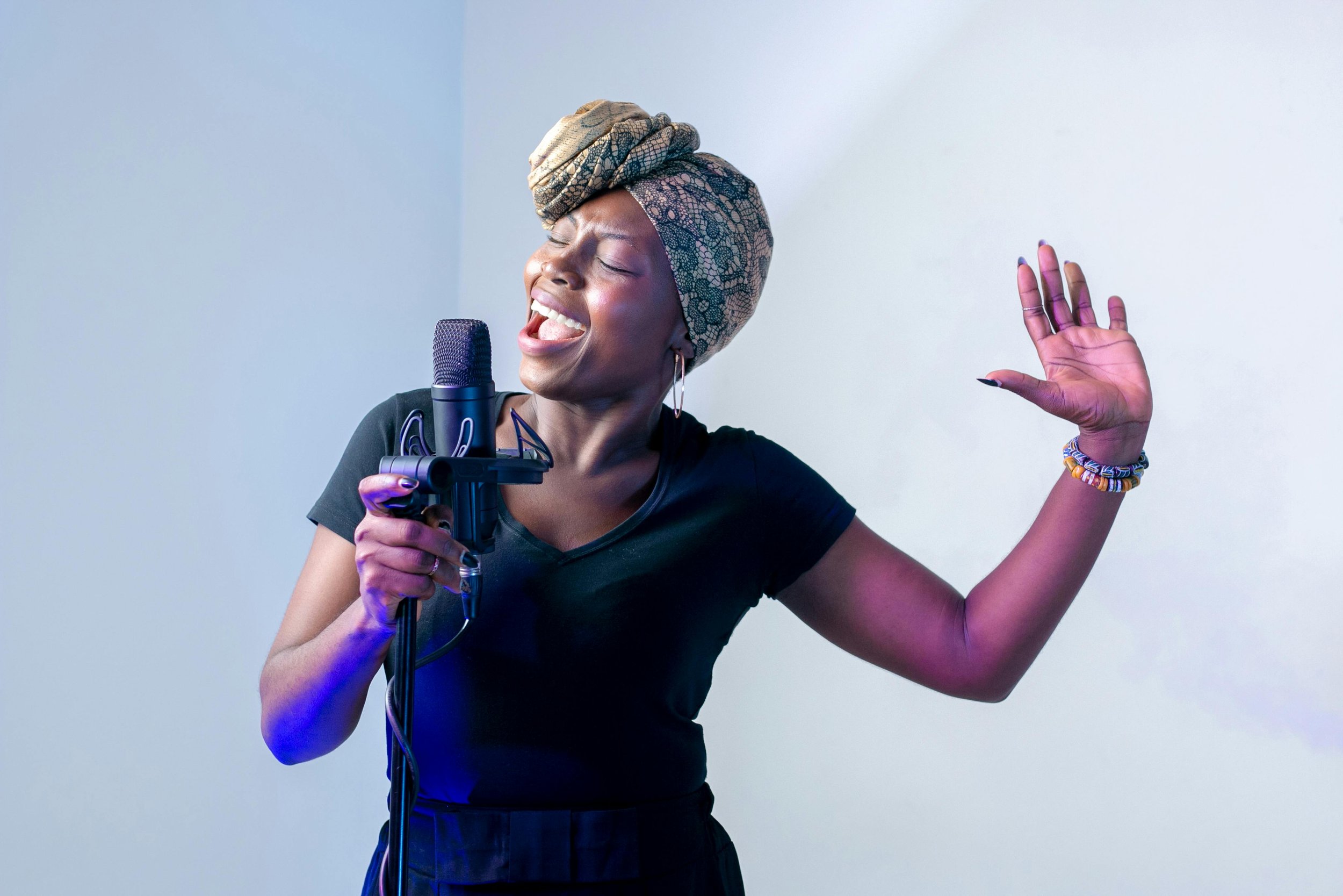 A woman singing passionately into a microphone with her eyes closed and one hand raised, wearing a patterned headwrap and casual black outfit.