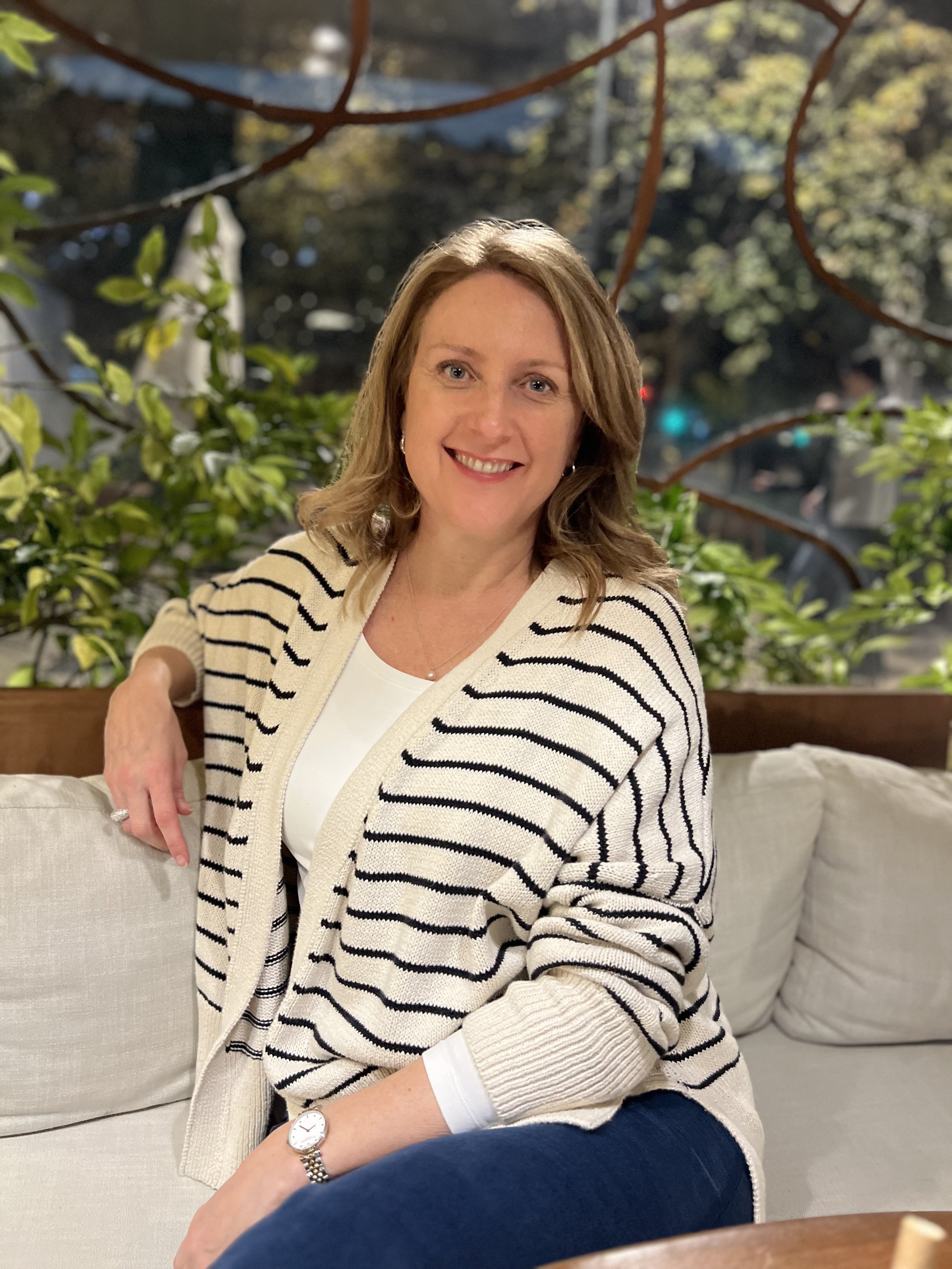 A casual image of Liz Flint wearing a white sweater with black stripes, is sitting on a cream-colored sofa in a warmly lit setting. She is smiling at the camera with a view of greenery and tree branches through the window behind her.