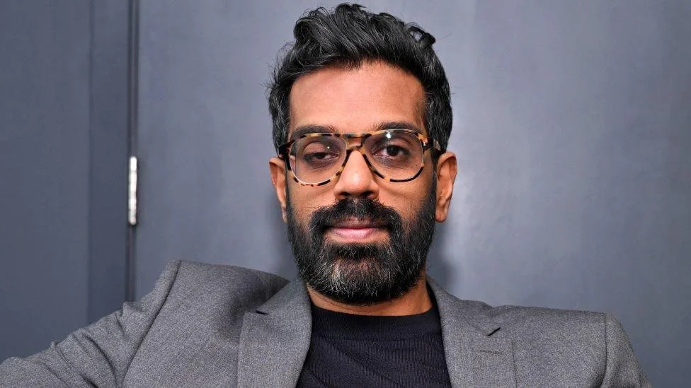 Romesh Ranganathan image with a beard, wearing glasses and a gray blazer, taking a selfie against a plain dark gray background.