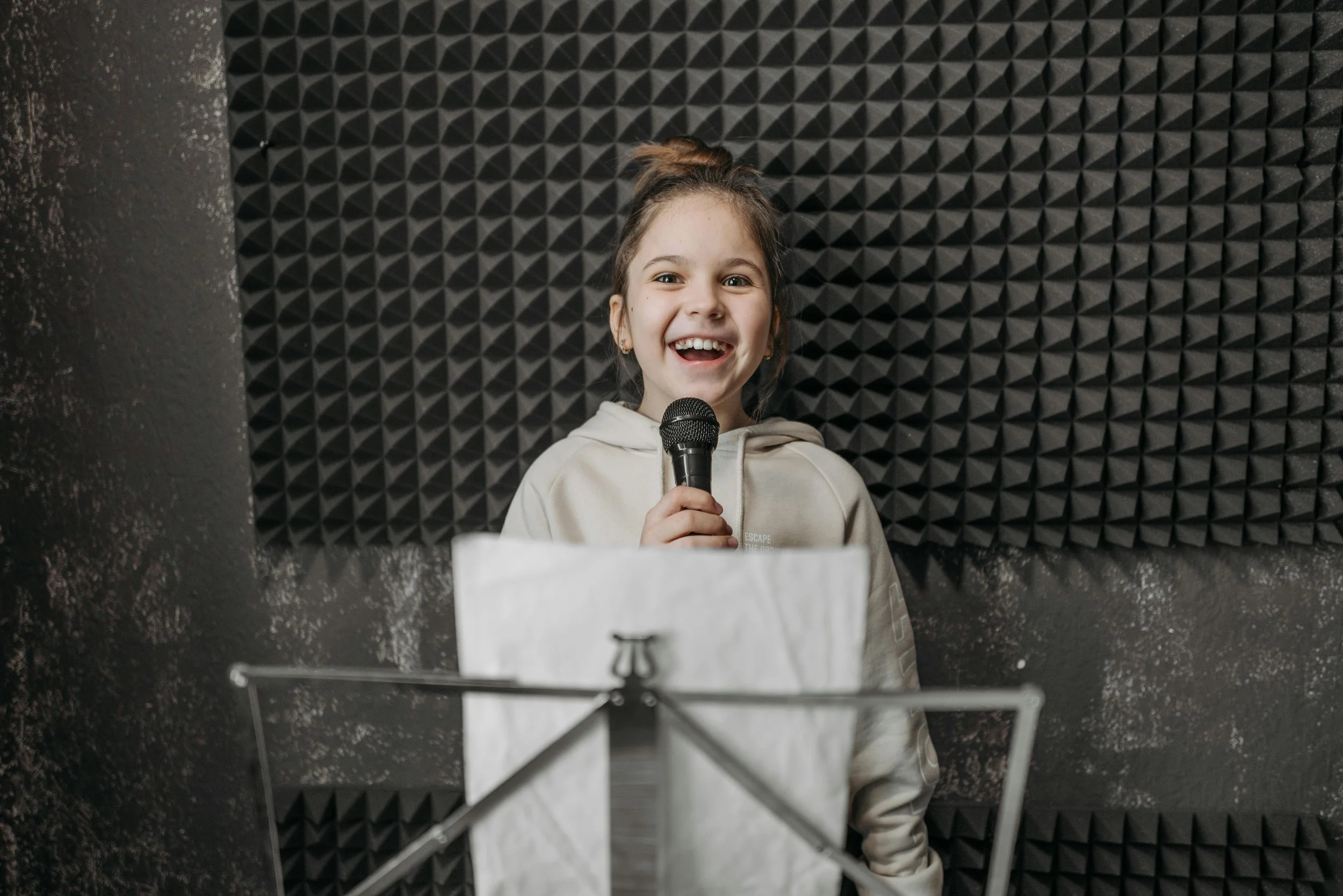 A young girl holding a microphone, smiling, in a recording studio with black acoustic foam panels on the wall.