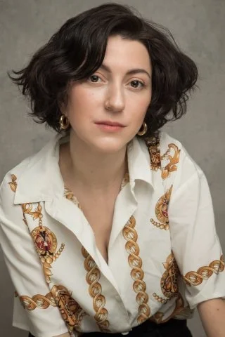 An image of Amalia Vitale with short wavy dark hair, wearing a white button-up shirt with gold chain patterns and gold hoop earrings, posing against a neutral background.