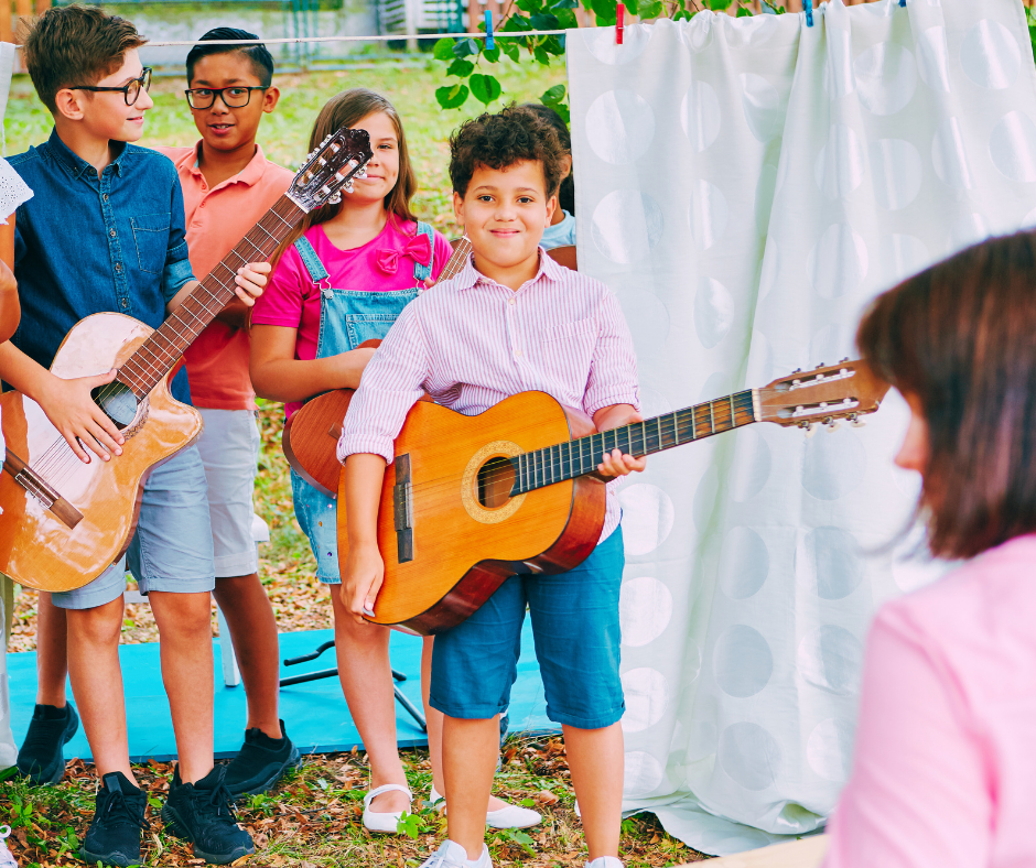 Children playing guitars outdoors during a performance or practice, with a woman watching and a white curtain backdrop.