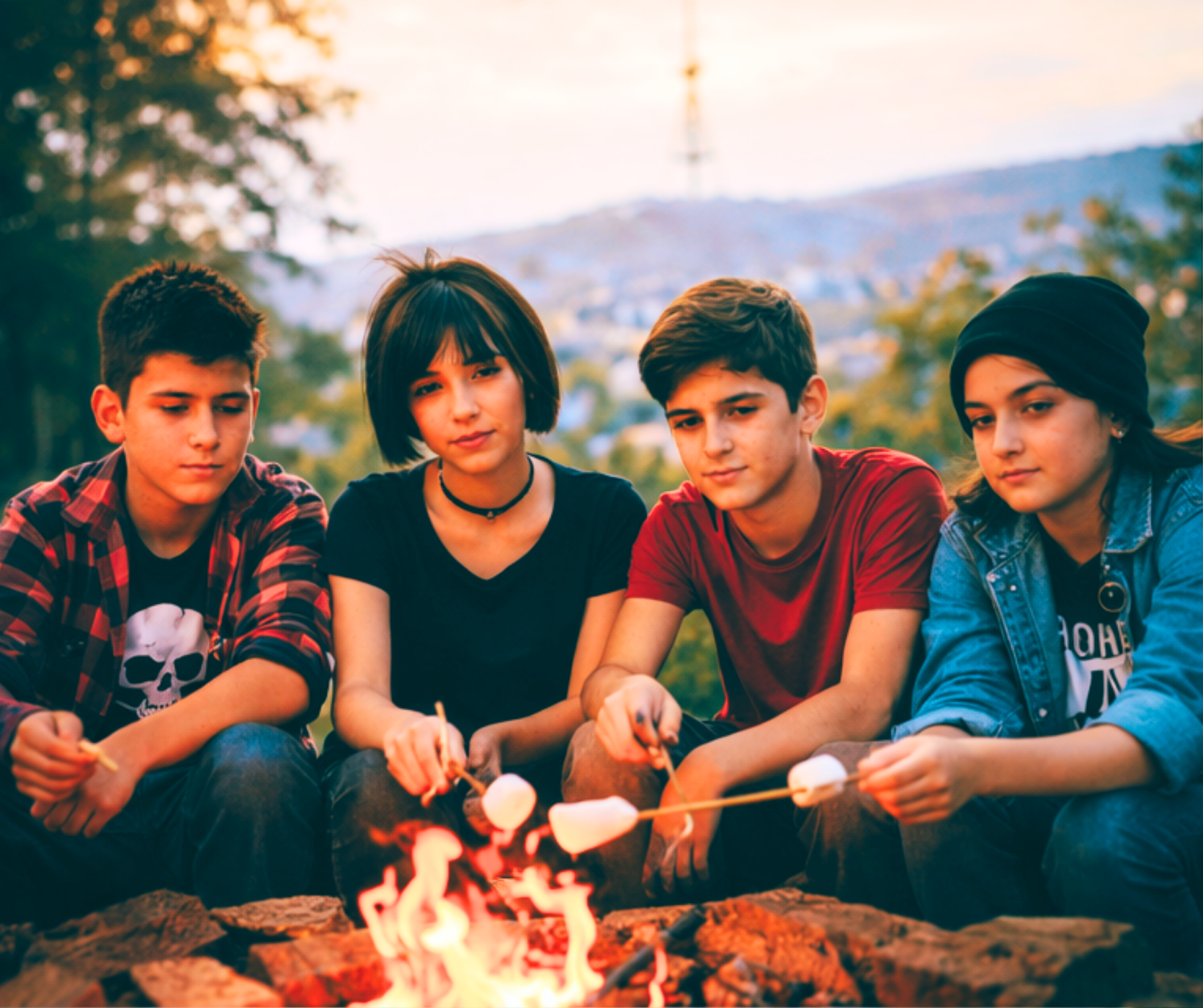 Four teenagers sitting around a campfire outdoors at sunset, roasting marshmallows.