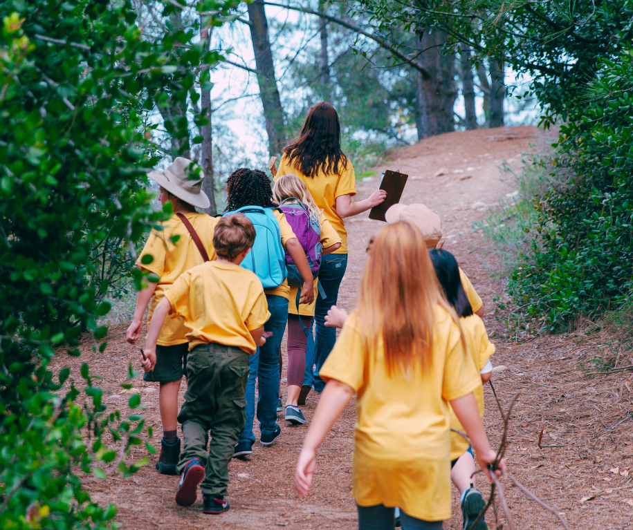 Children and a young adult hiking through a wooded trail, some carrying backpacks and wearing yellow shirts.