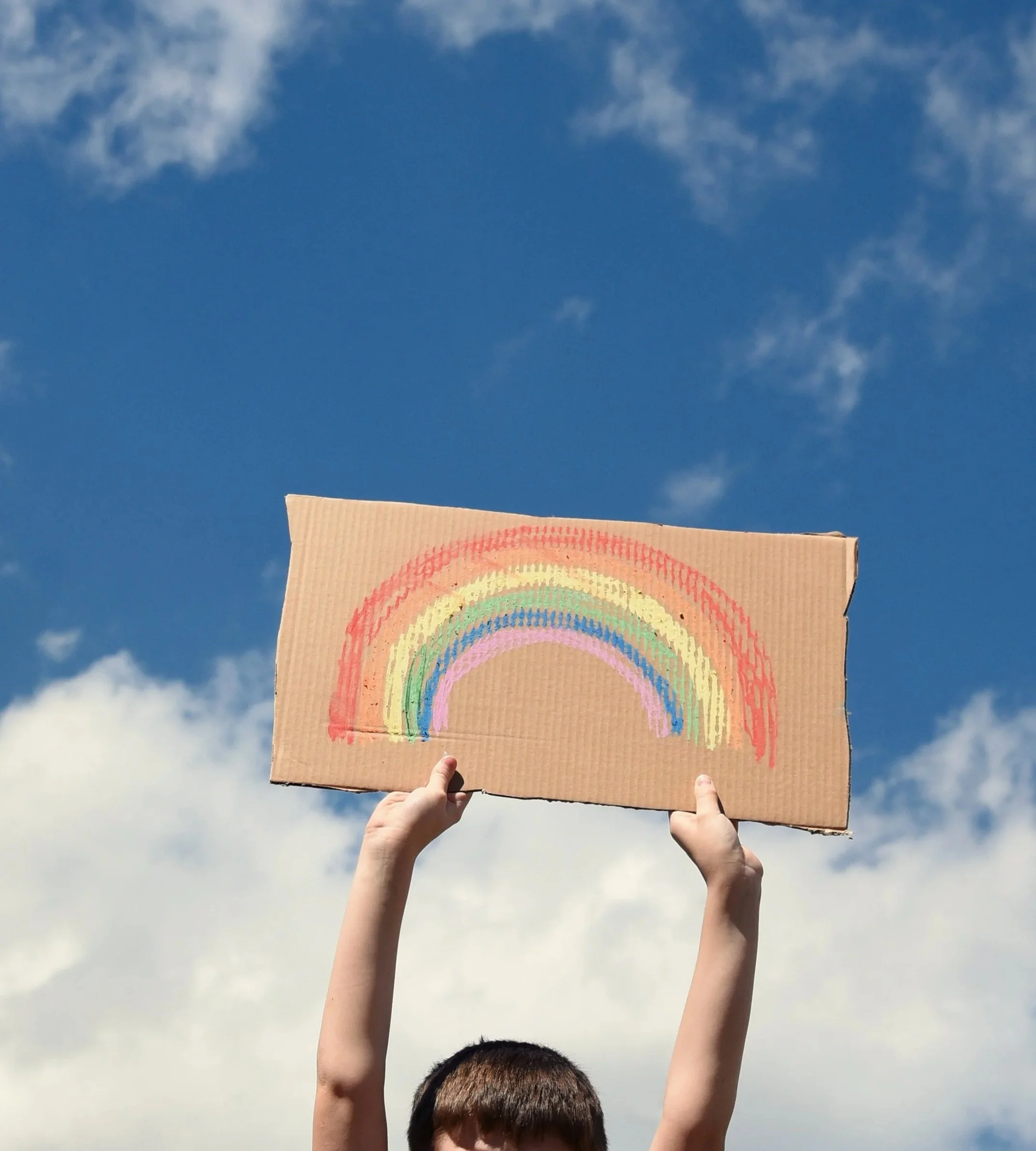 Person holding up a cardboard sign with a rainbow painted on it against a blue sky with clouds.