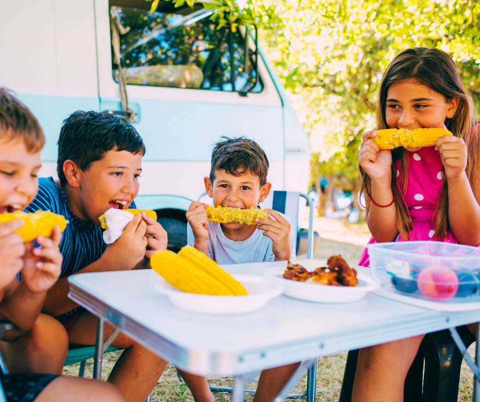 Kids enjoying corn on the cob at an outdoor gathering with food on a table at a park.