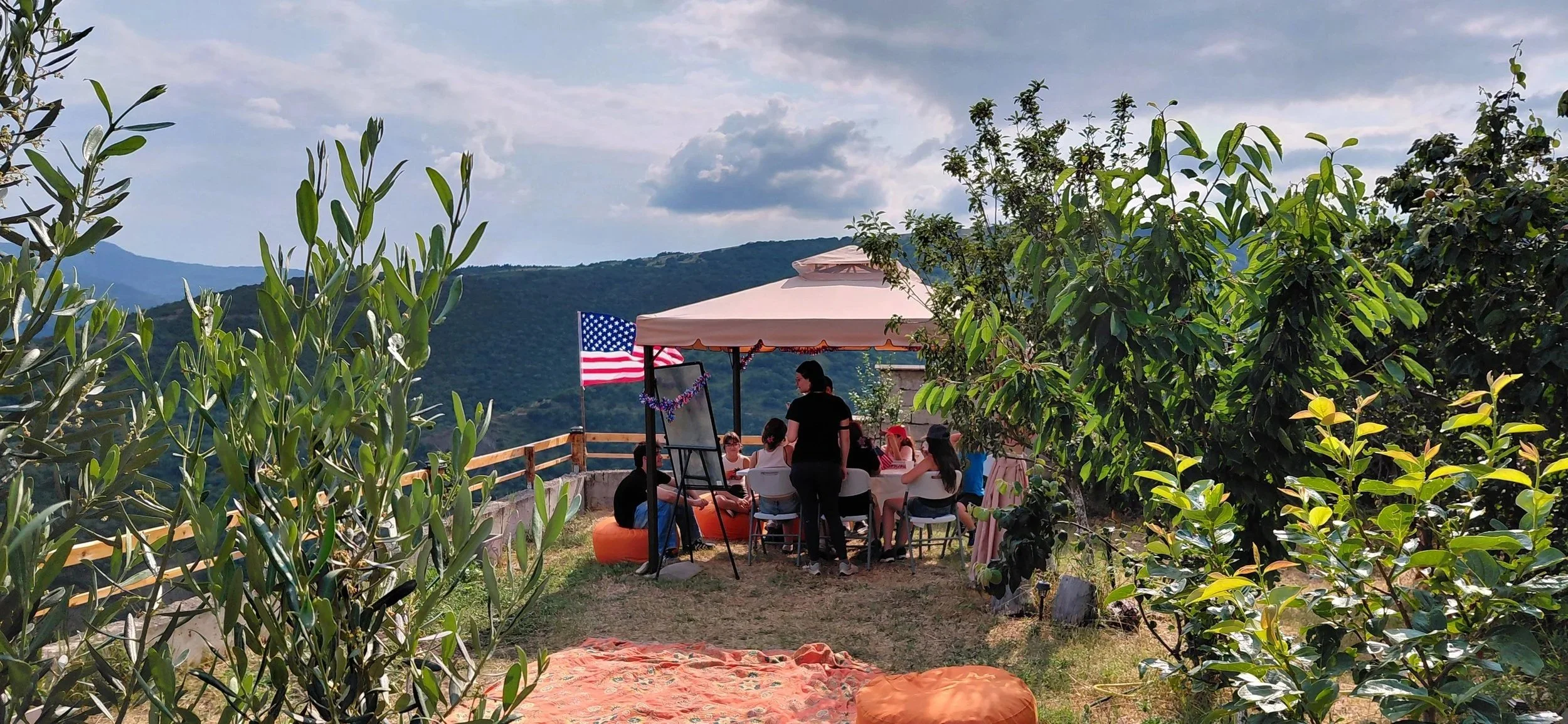 People gathered under a small canopy with an American flag, sitting around a table on a mountain terrace surrounded by greenery and trees, with mountains in the background.