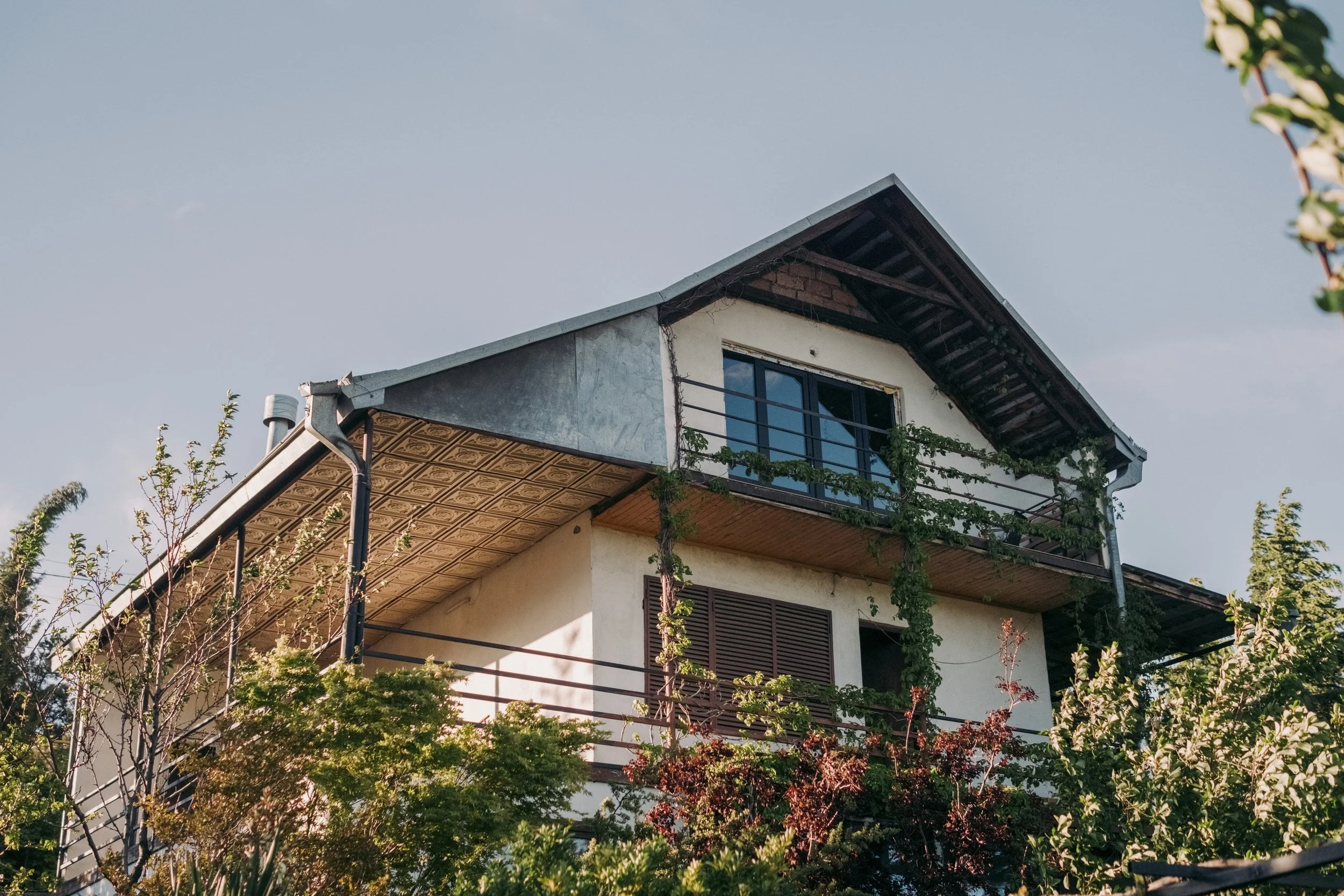 Napo Garden House , the home of Camp Americana - a modern house with a sloped roof, featuring a balcony with a metal railing and large glass doors, surrounded by green trees and shrubs under a clear sky.