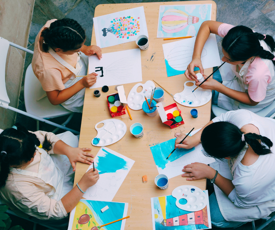 Four young girls are seated around a wooden table engaging in painting and drawing activities. They have various art supplies, including paint palettes, brushes, and cups of water. The table features colorful artwork, including a picture of a hot air balloon, a cluster of balloons, and other watercolor paintings.