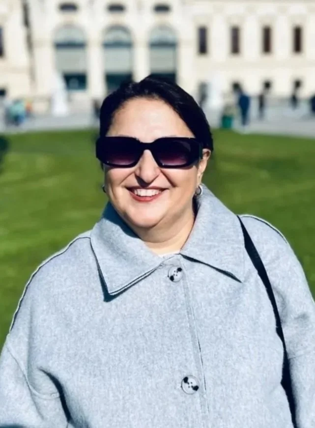 A smiling woman with dark hair wearing large sunglasses and a gray coat, standing outdoors on a sunny day with a building and grassy area in the background.