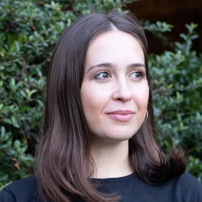 A young woman with long brown hair, wearing a black shirt, standing outdoors in front of green bushes.