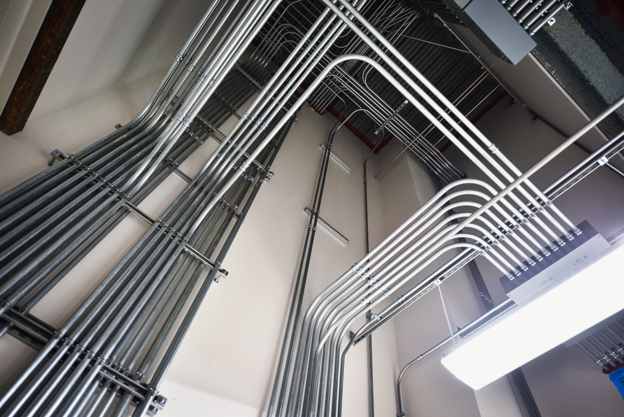 A room with organized electrical conduit pipes running along the ceiling and wall, supported by metal brackets and labeled with tags.