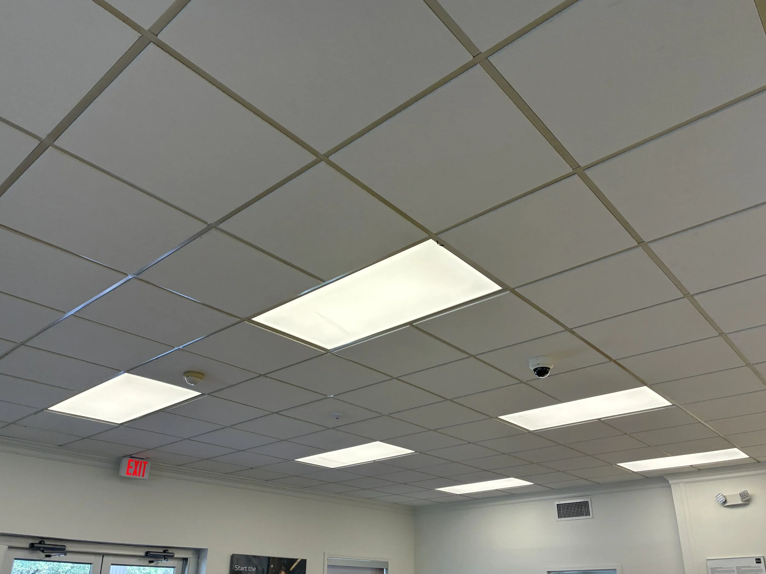 Ceiling with rectangular fluorescent lights in a room, an exit sign, a security camera, and a wall vent.