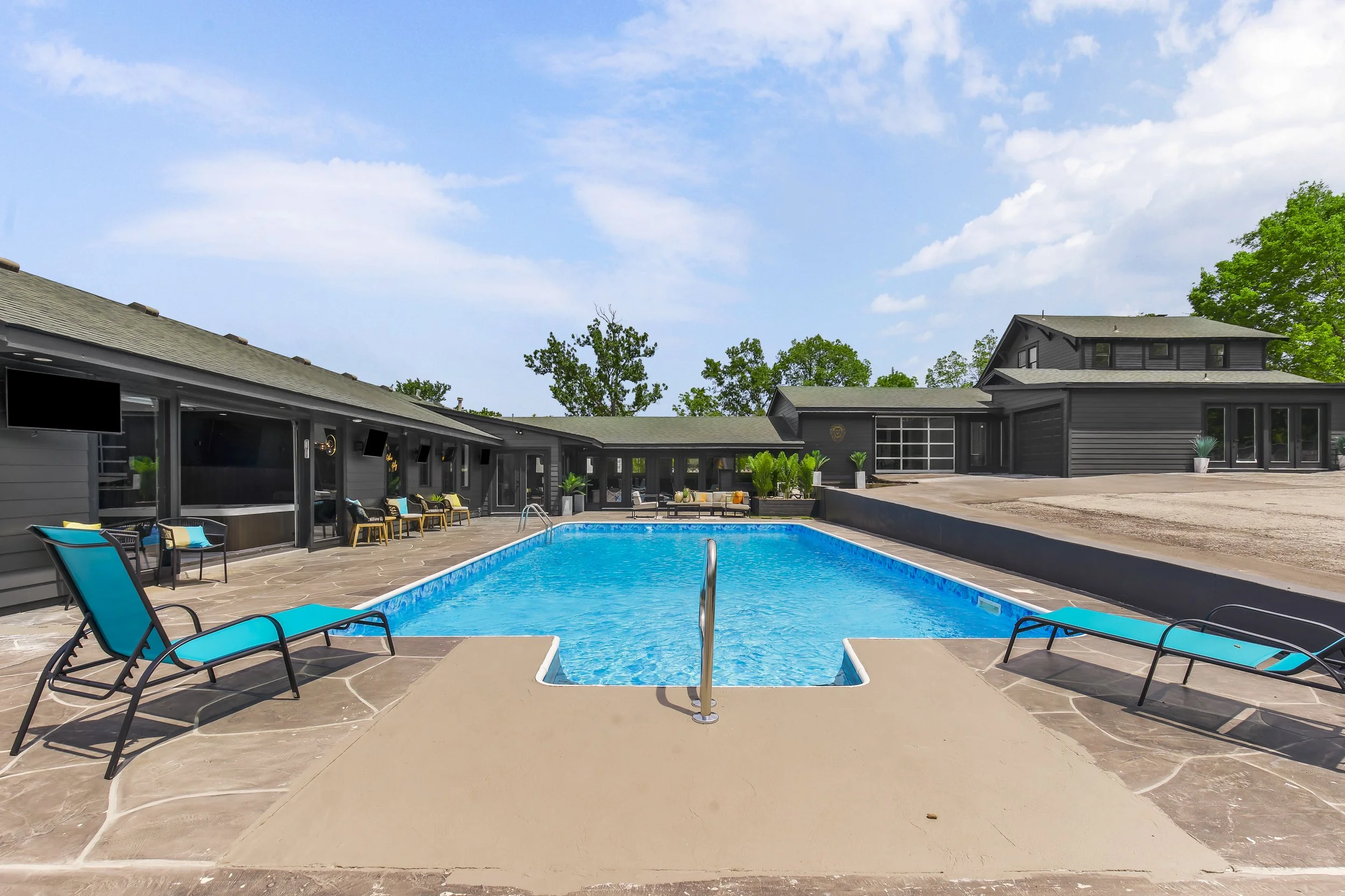 A modern house with a backyard pool, lounge chairs, and surrounding patio furniture under a blue sky with clouds.