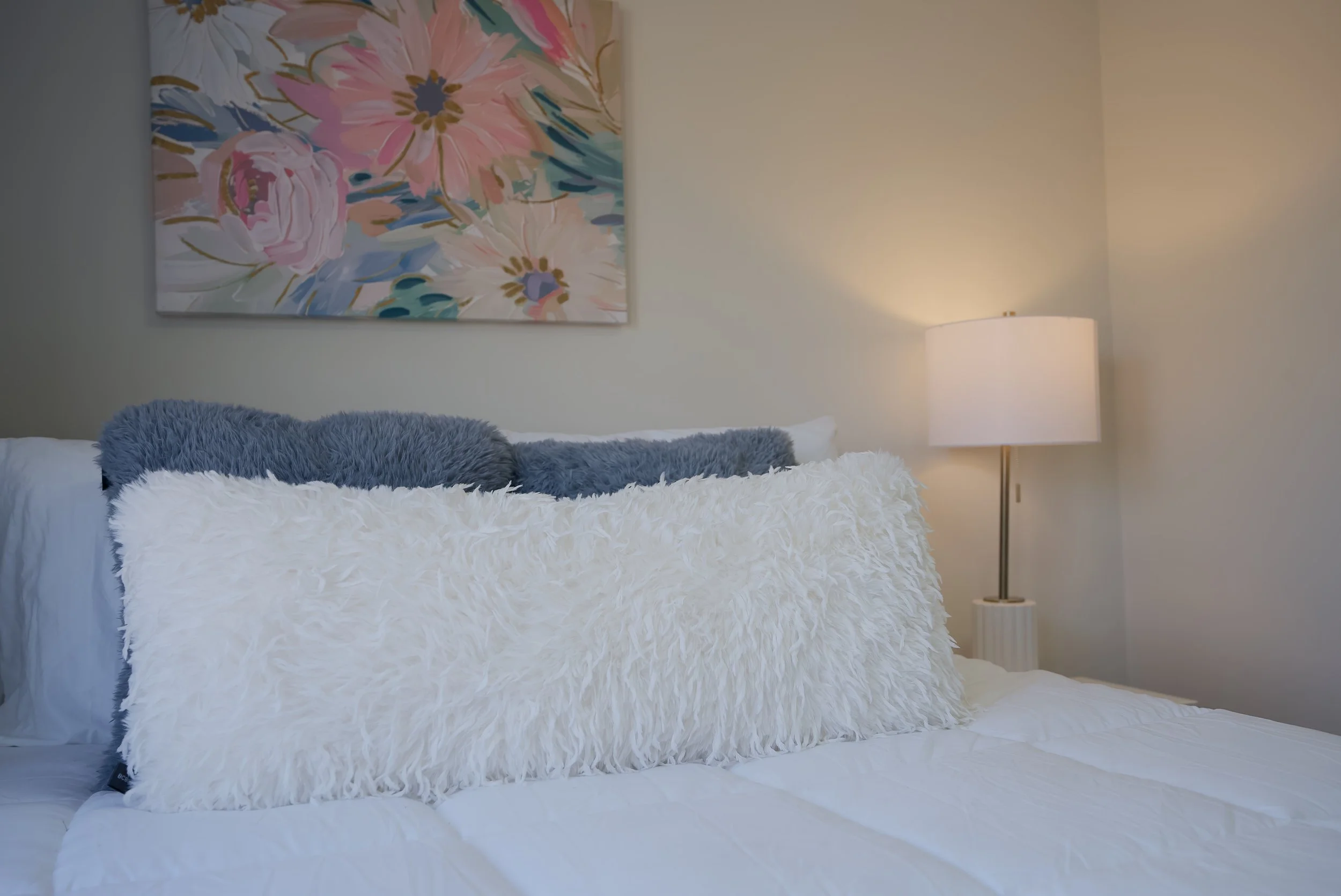 A neatly made bed with a fluffy white pillow and a smaller gray pillow in a bedroom. Above the bed, there is a colorful floral painting. To the right, a standing lamp emits warm light against the plain wall.
