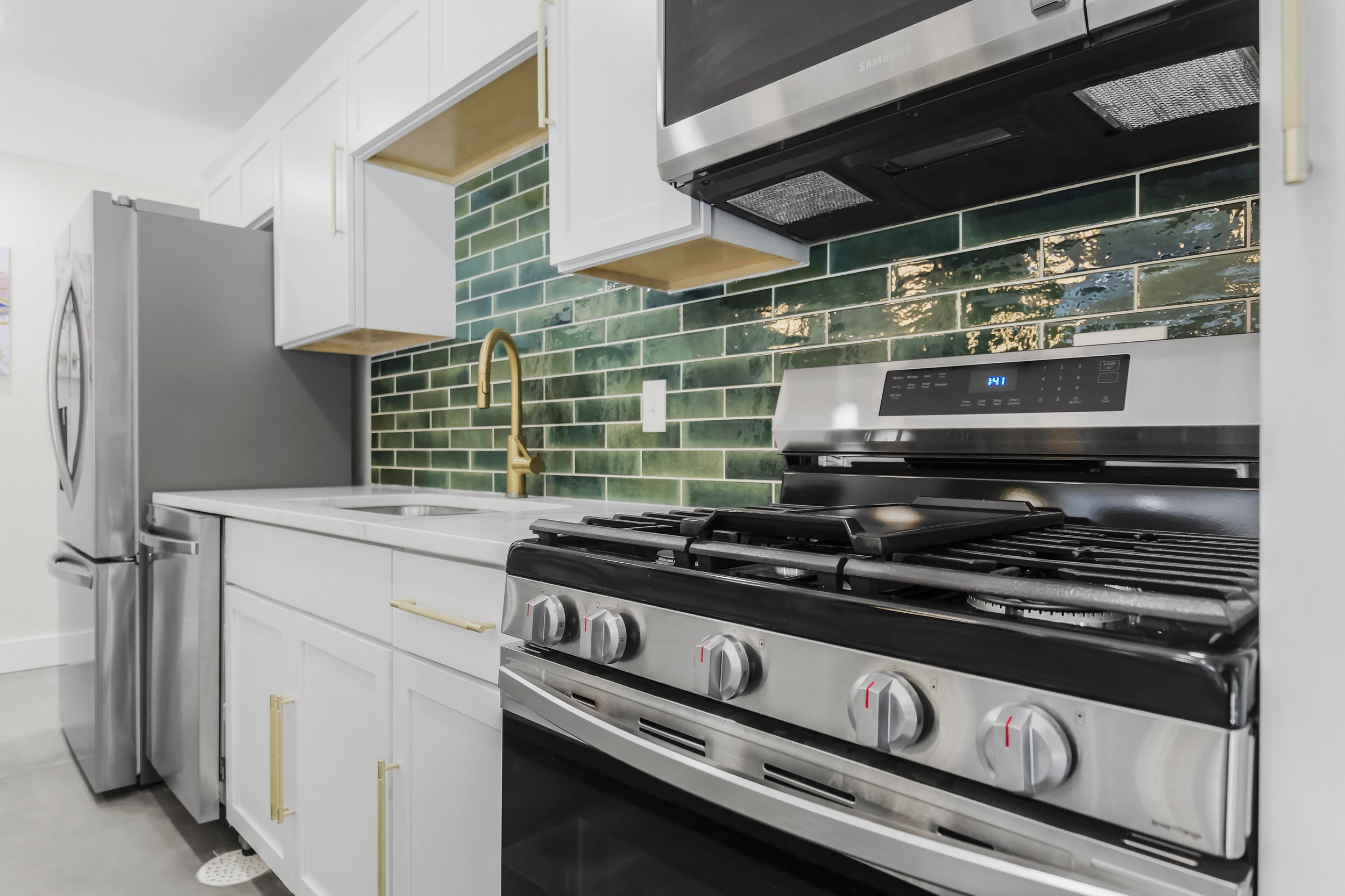 Modern kitchen with green tiled backsplash, white cabinets, stainless steel refrigerator, oven, microwave, gold faucet, and white countertop.