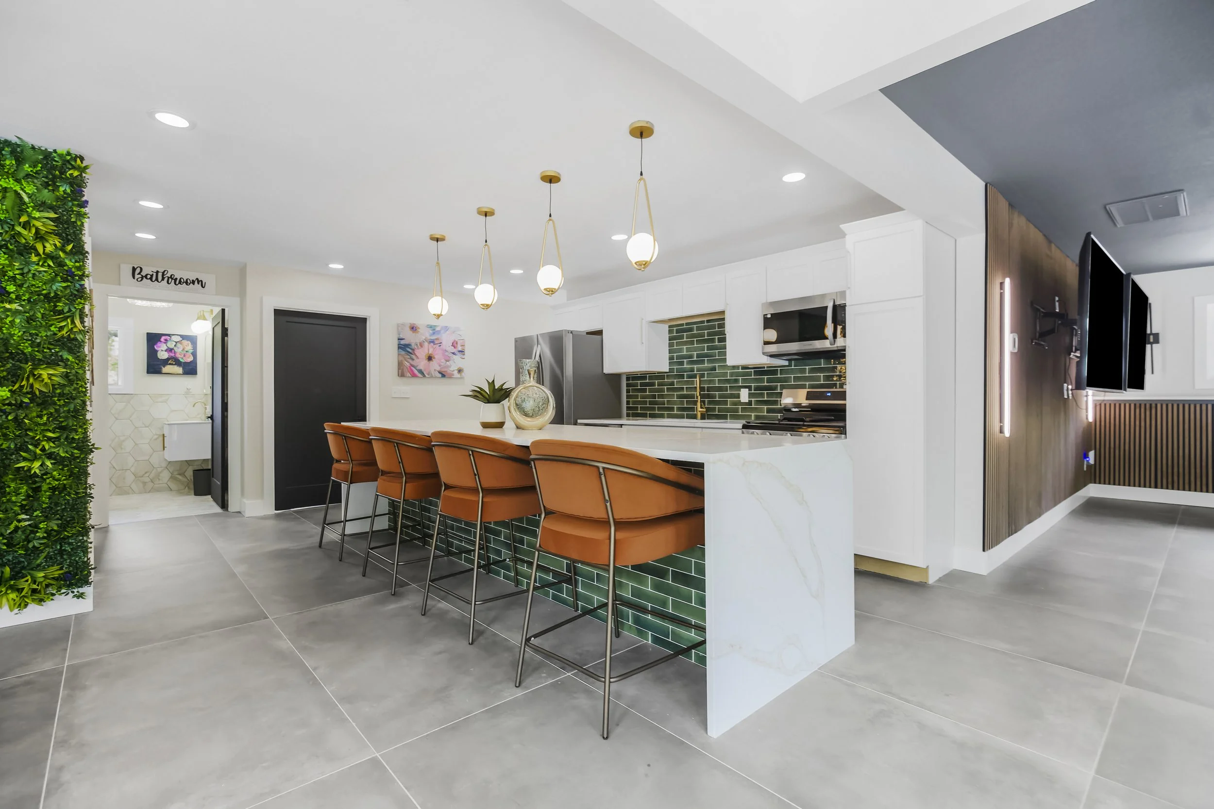 Modern kitchen with white cabinetry, green tile backsplash, marble island, four brown barstools, pendant lighting, and a living area with a wall-mounted TV.