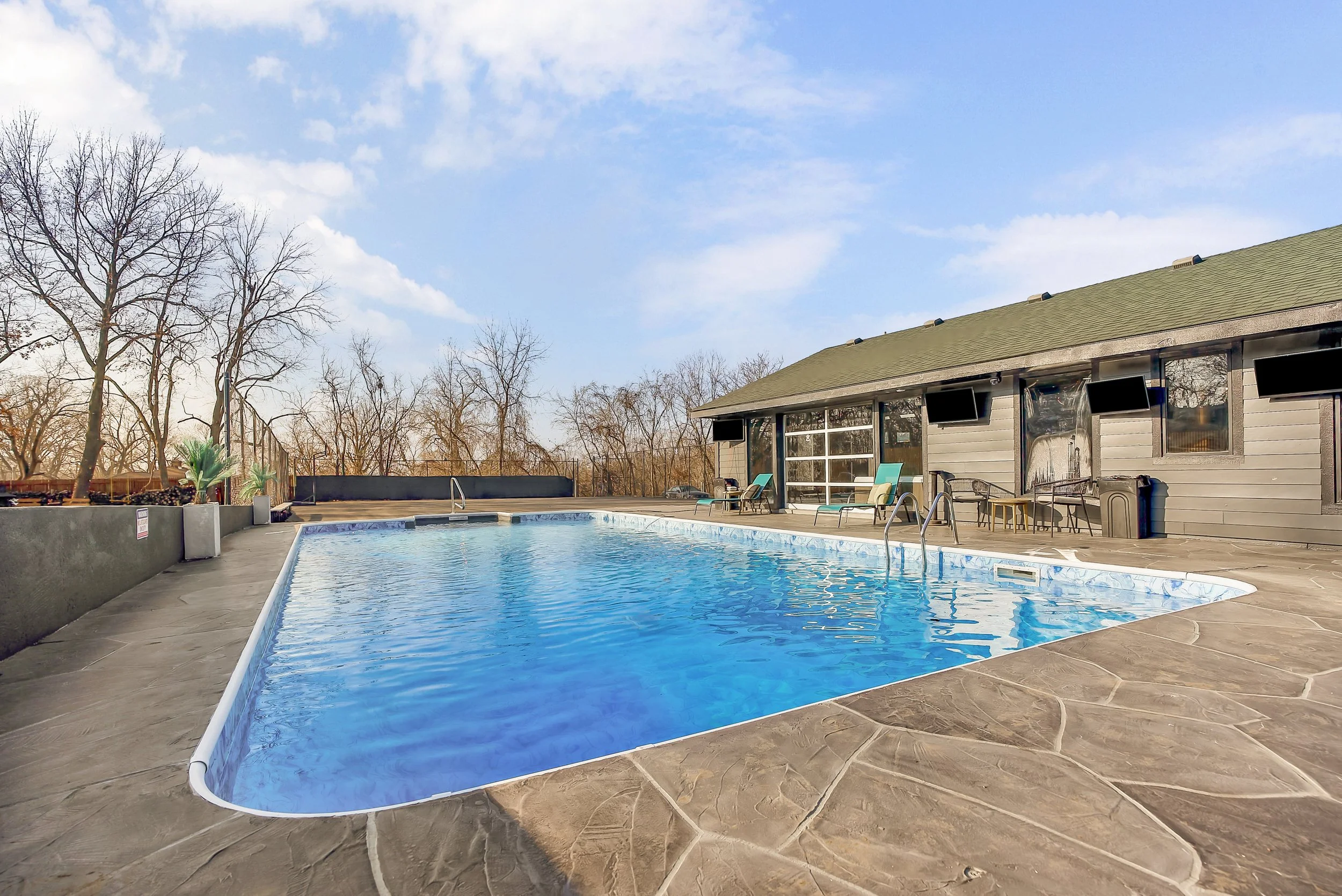 An outdoor swimming pool with a clear blue sky, leafless trees, and a house with a porch and outdoor chairs in the background.