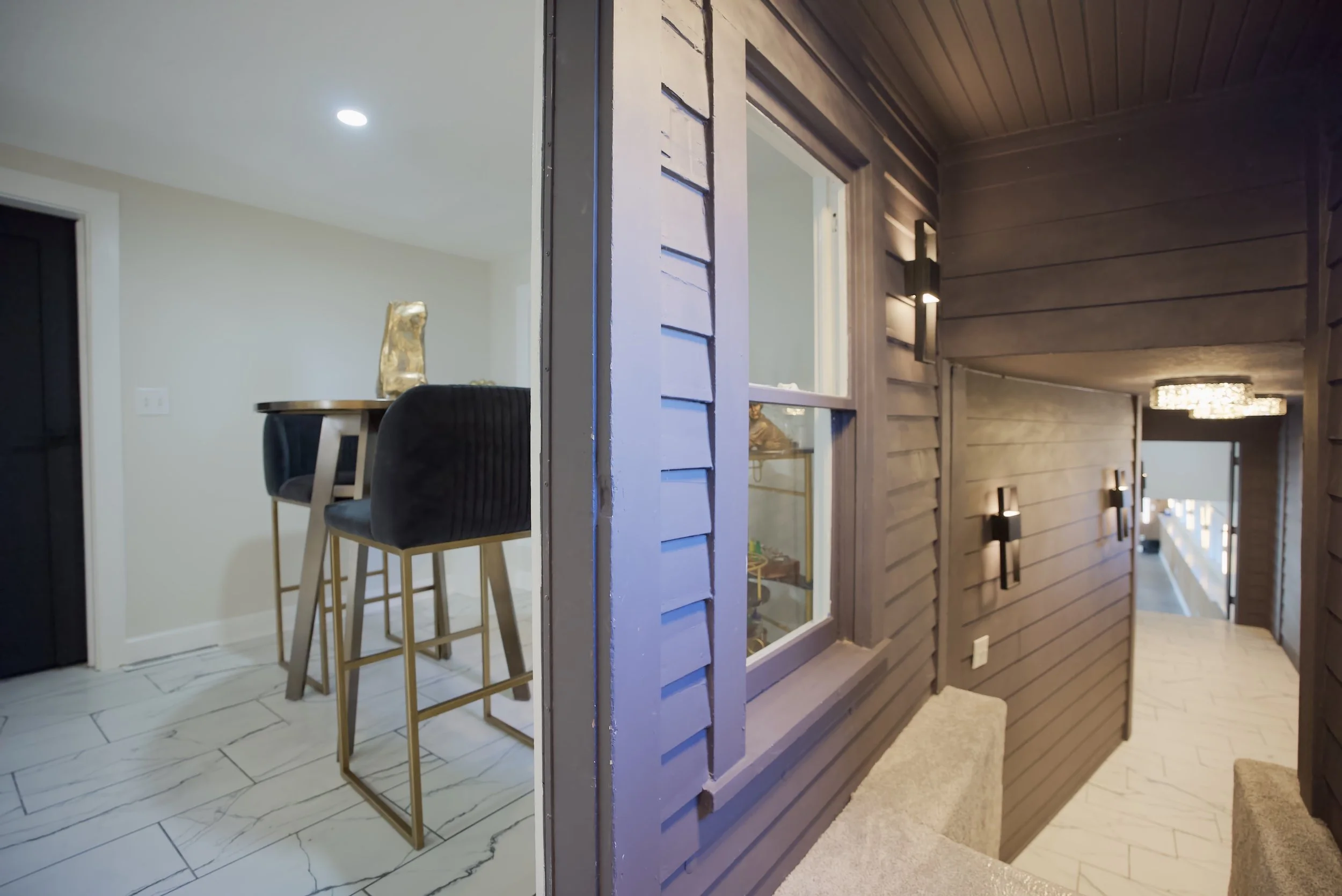 A split image showing a dining area with marble floor tiles and two black velvet chairs at a high wooden table on the left, and a hallway with brown wooden paneling on the right, featuring wall-mounted lights and a staircase at the end.