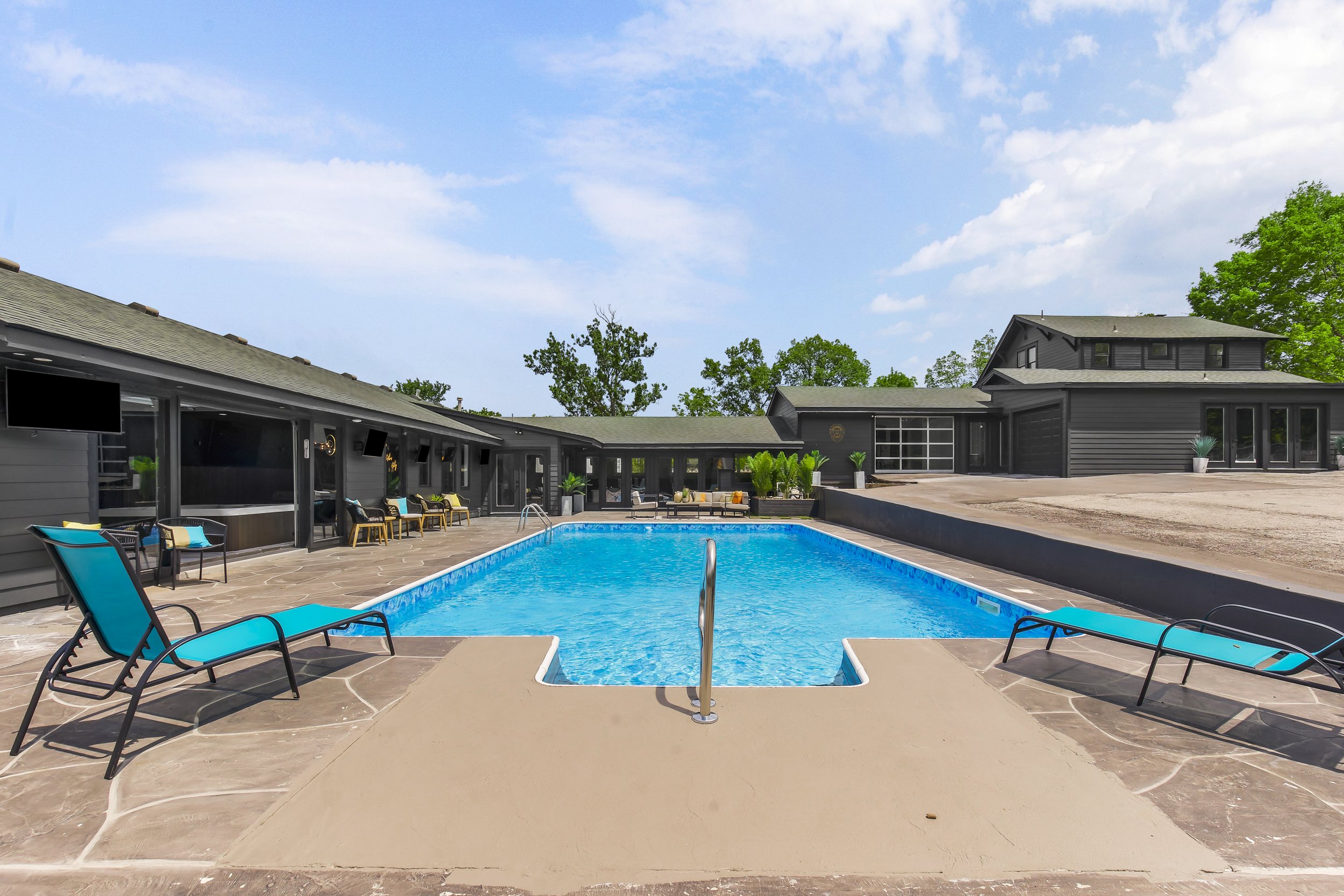 A backyard swimming pool with blue water, surrounded by a patio with lounge chairs and seating areas. There are black houses or buildings with windows in the background and a partly cloudy sky above.