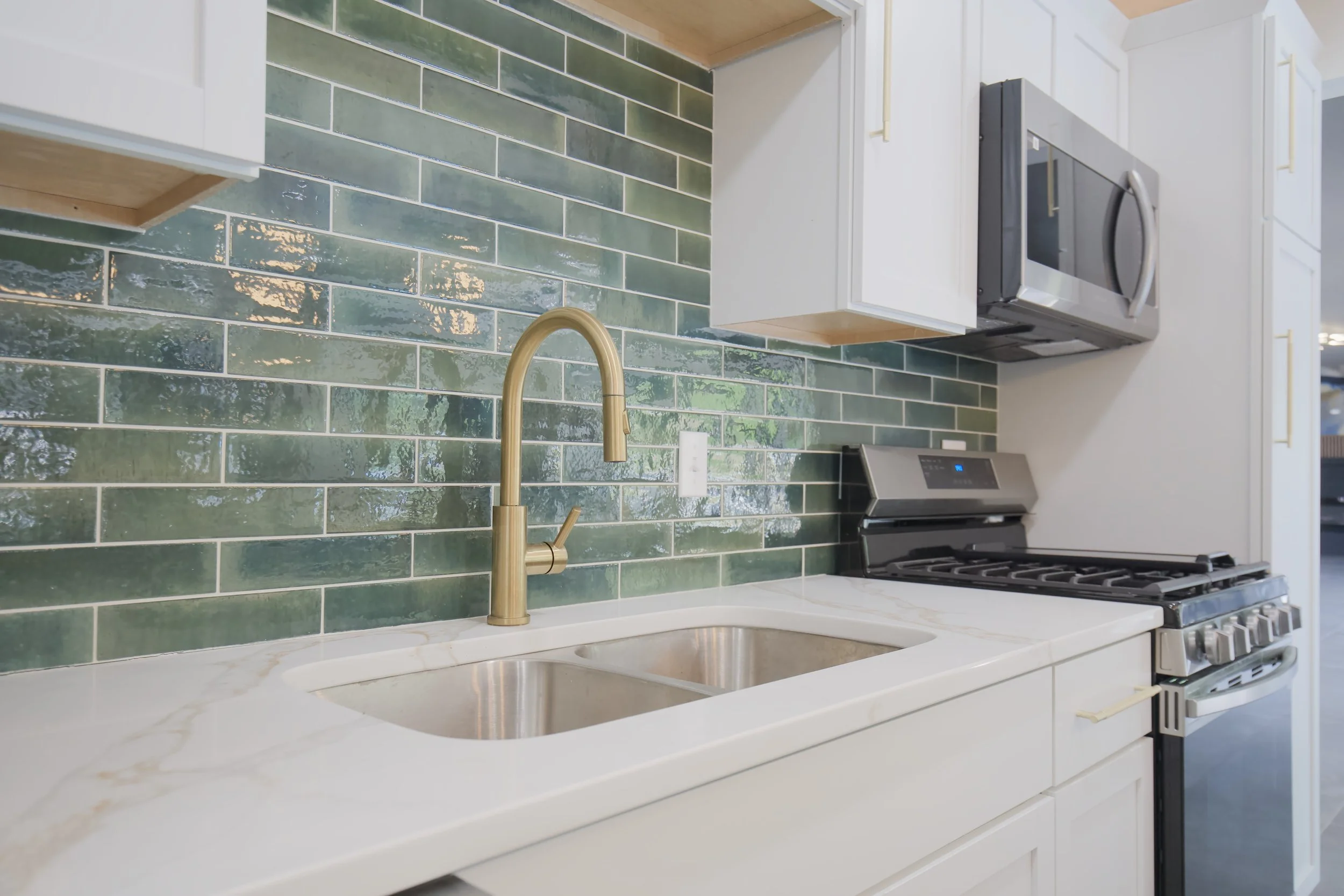 Kitchen with green tiled backsplash, white countertop with a double sink, brass faucet, black and stainless steel stove, microwave, and white cabinets.