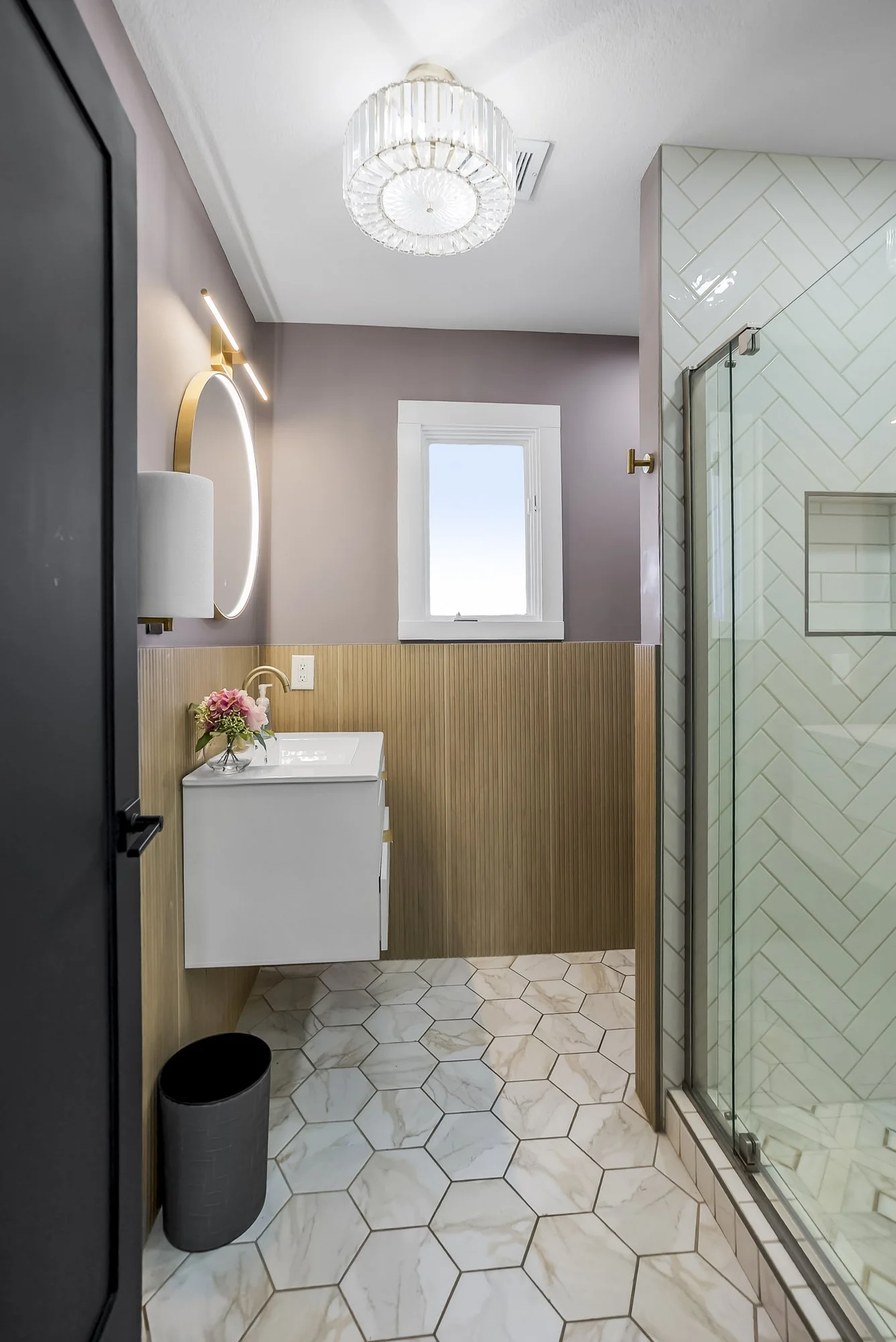 Modern bathroom with hexagon marble floor tiles, a small floating white vanity with a gold faucet, a round mirror with built-in lighting, a window with white trim, a glass shower with white herringbone tiles, and a decorative chandelier ceiling light