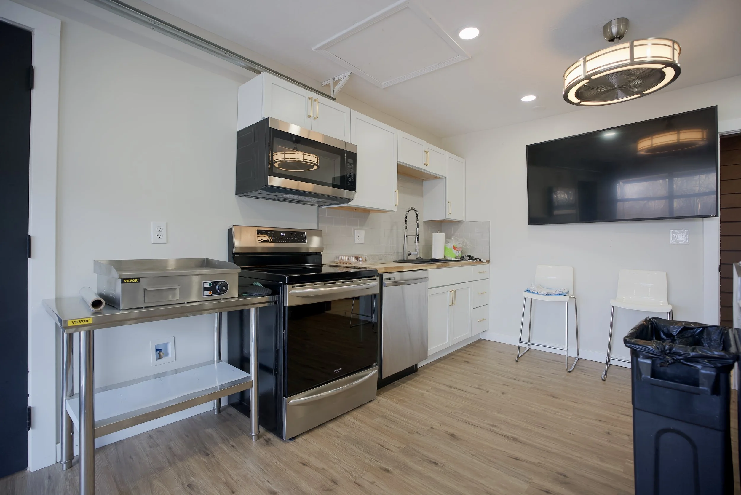 Kitchen with white cabinets, stainless steel appliances, a microwave above the stove, a sink, and a large flat-screen TV mounted on the wall. There are two white chairs, a trash bin, and a cart with a food warmer.