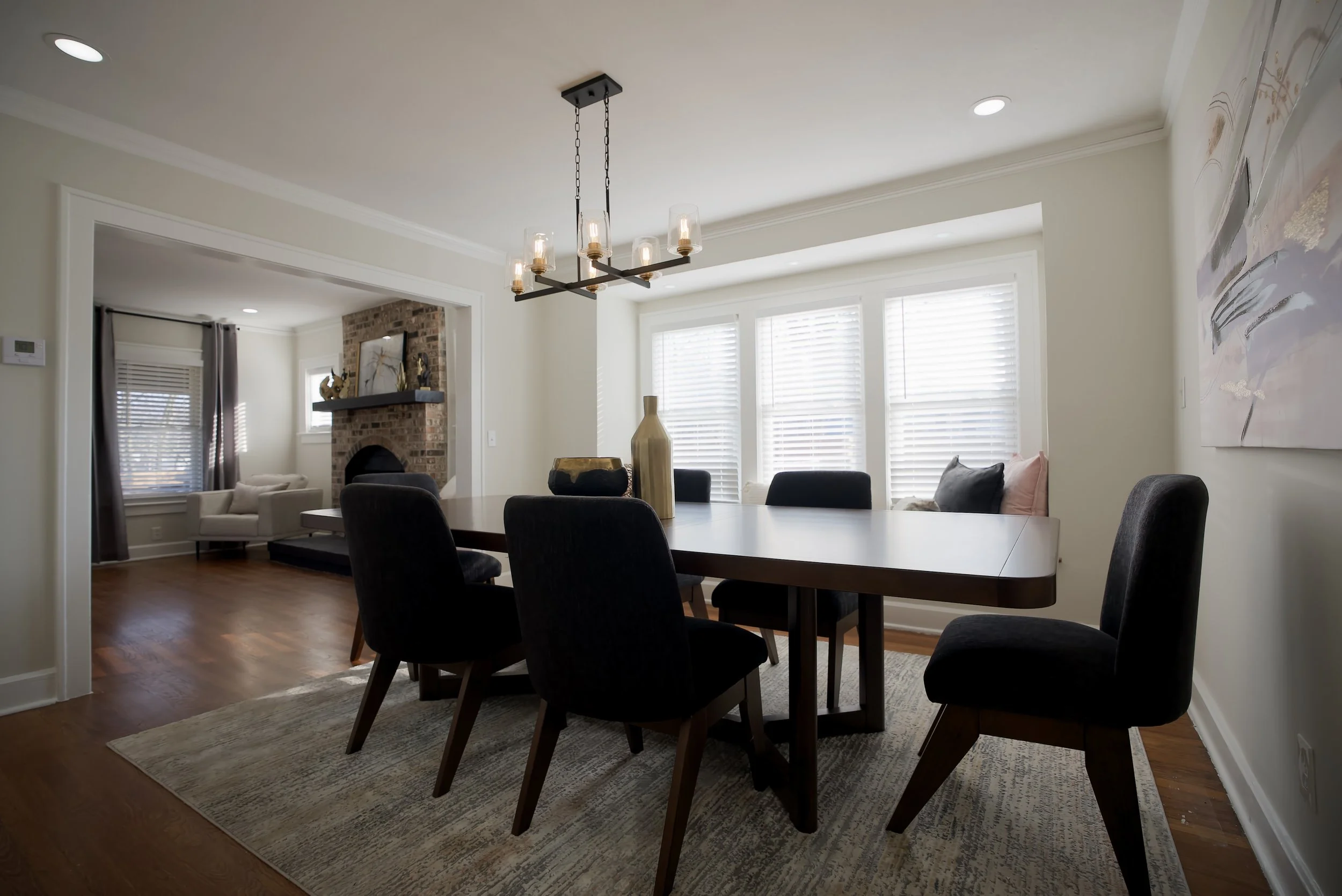 Dining room with a wooden table, six black chairs, a chandelier with multiple exposed bulbs, decorative vases on the table, large window with white blinds, and a living room with a fireplace and armchair visible in the background.