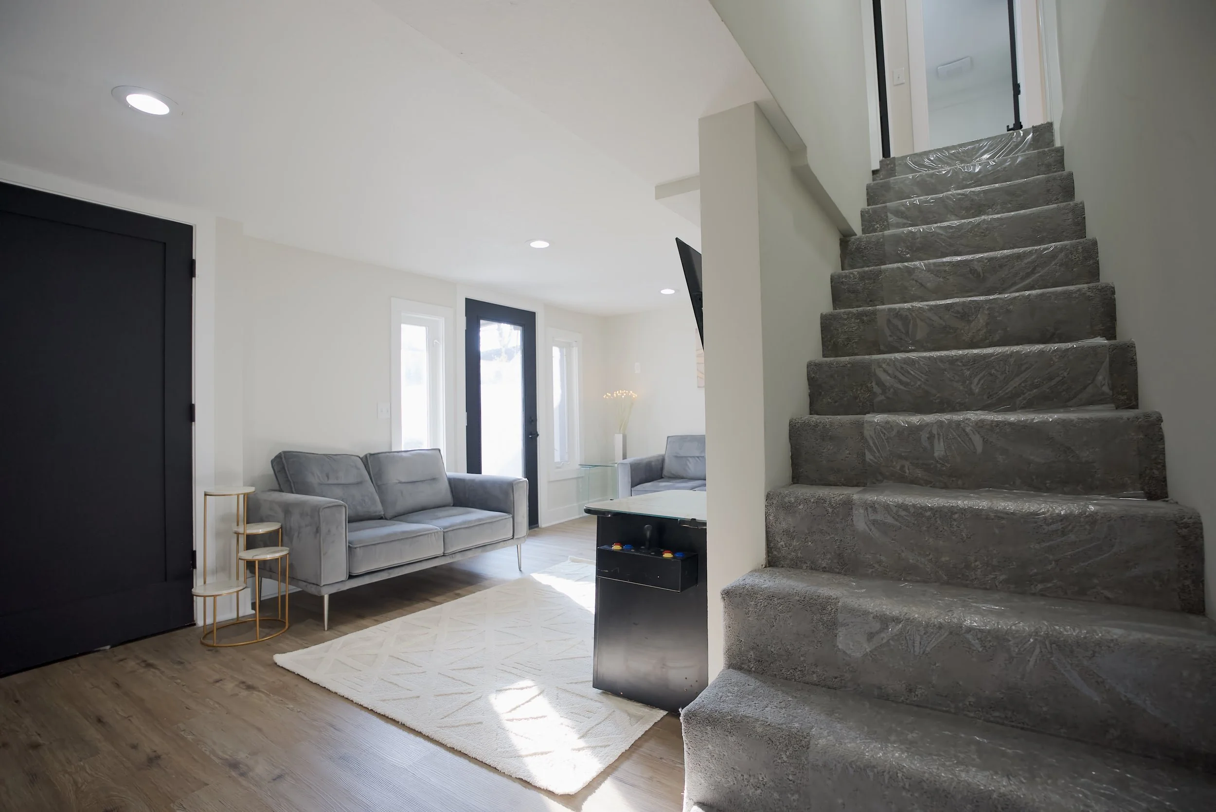 Living room with gray sofas, a white rug, and a glass-top table, adjacent to a staircase with gray marble steps and plastic wrapping. Natural light coming through windows and a door.