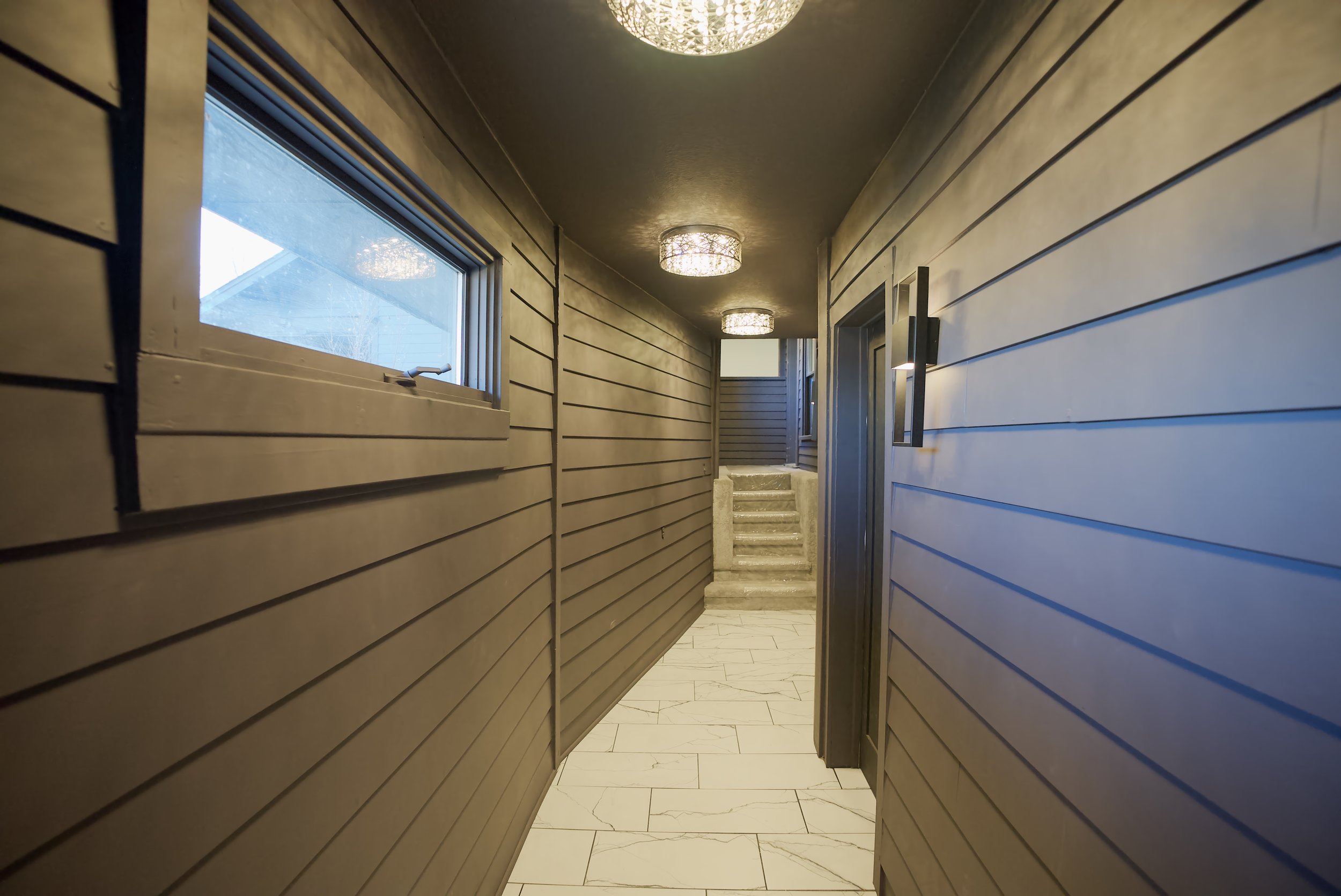 Indoor corridor with brown horizontal wooden panels, a window on the left, modern ceiling lights, and a short staircase at the end.