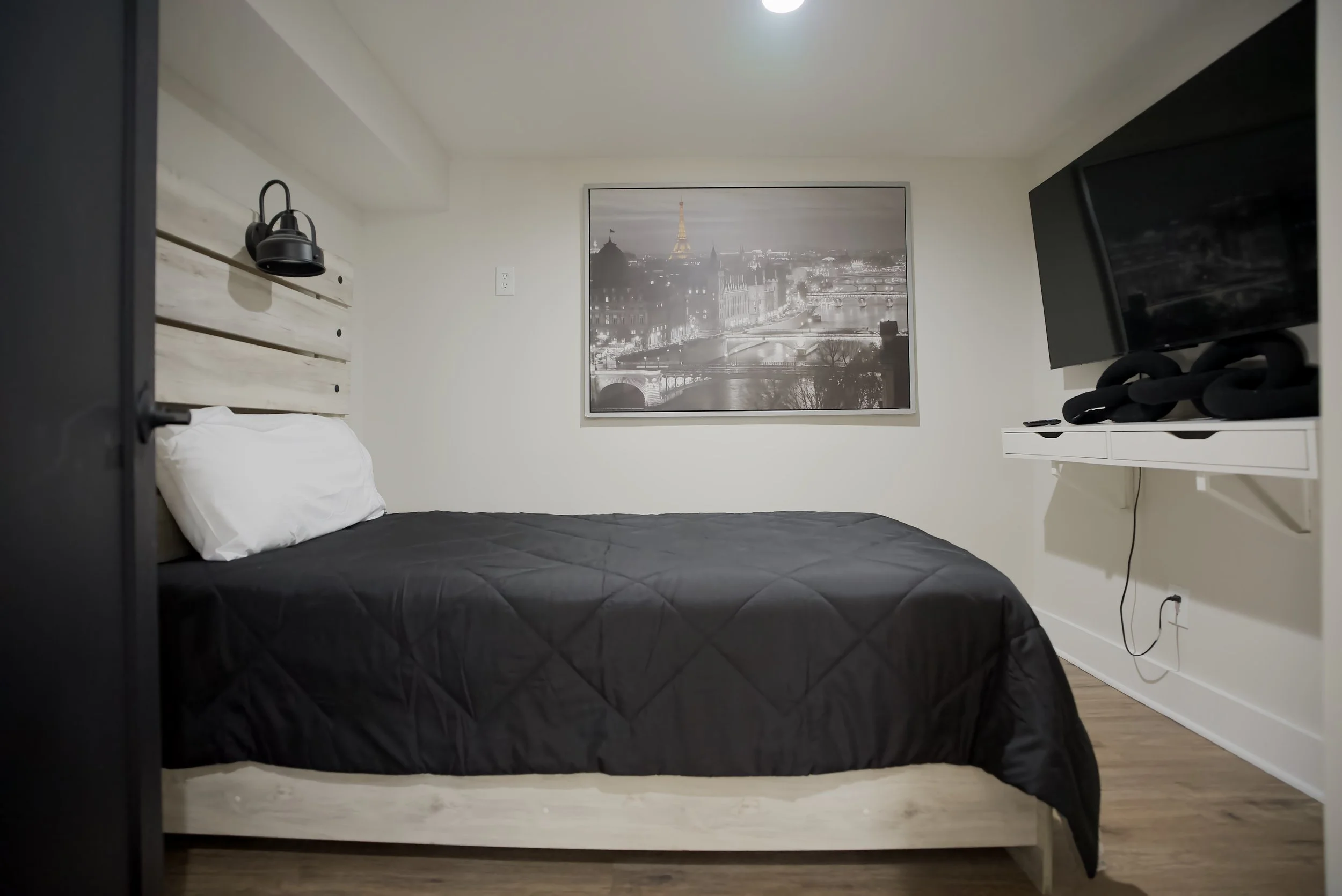 A minimalist bedroom with a bed covered in a black quilt, a white pillow, a wooden headboard, a wall-mounted black lamp, a large picture of Paris with the Eiffel Tower, and a wall-mounted TV on a white shelf.