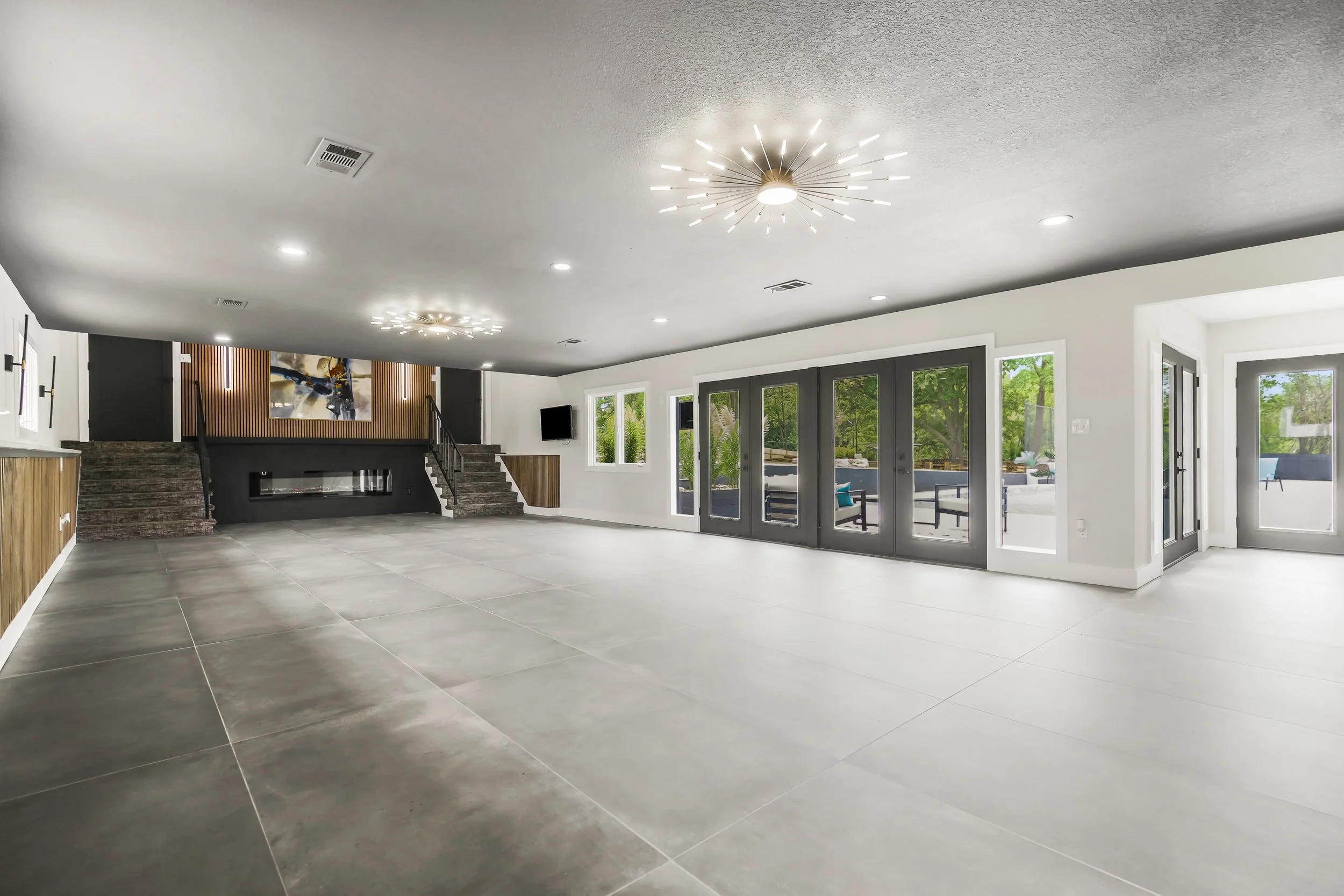 Empty modern indoor room with large tiled floor, white walls, multiple ceiling lights, and glass doors leading outside, with greenery visible through the windows.