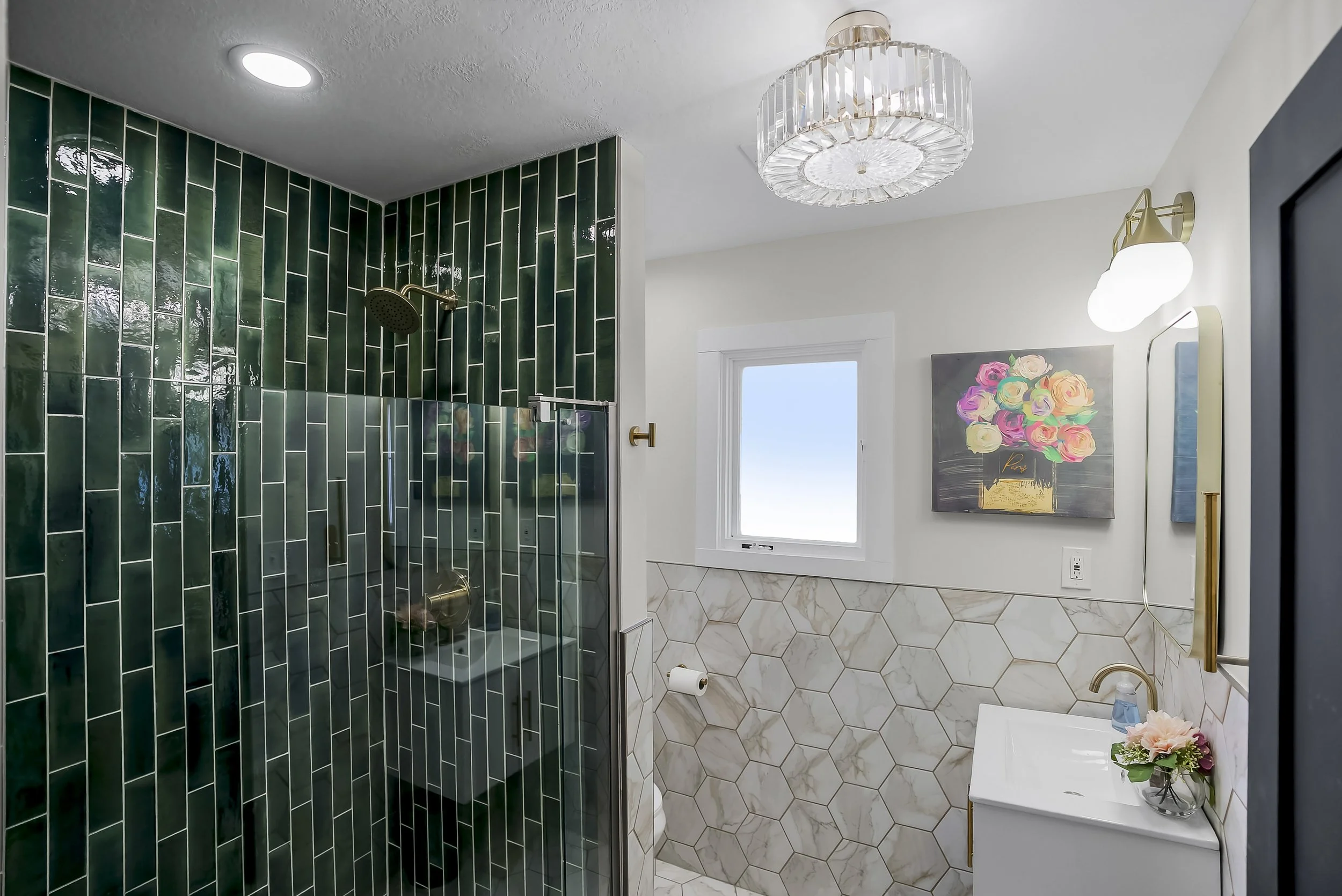 Bathroom with green tiled shower, white vanity with flowers, wall art of roses, window, and modern lighting fixtures.