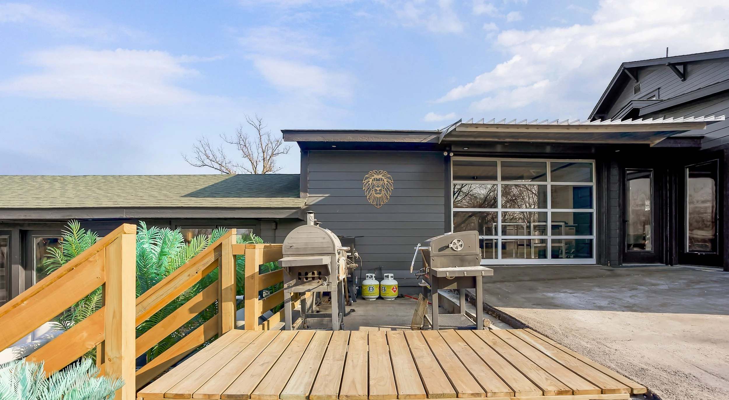 Modern residential backyard with a wooden deck, outdoor grills, propane tanks, and a gray exterior house with large glass windows.