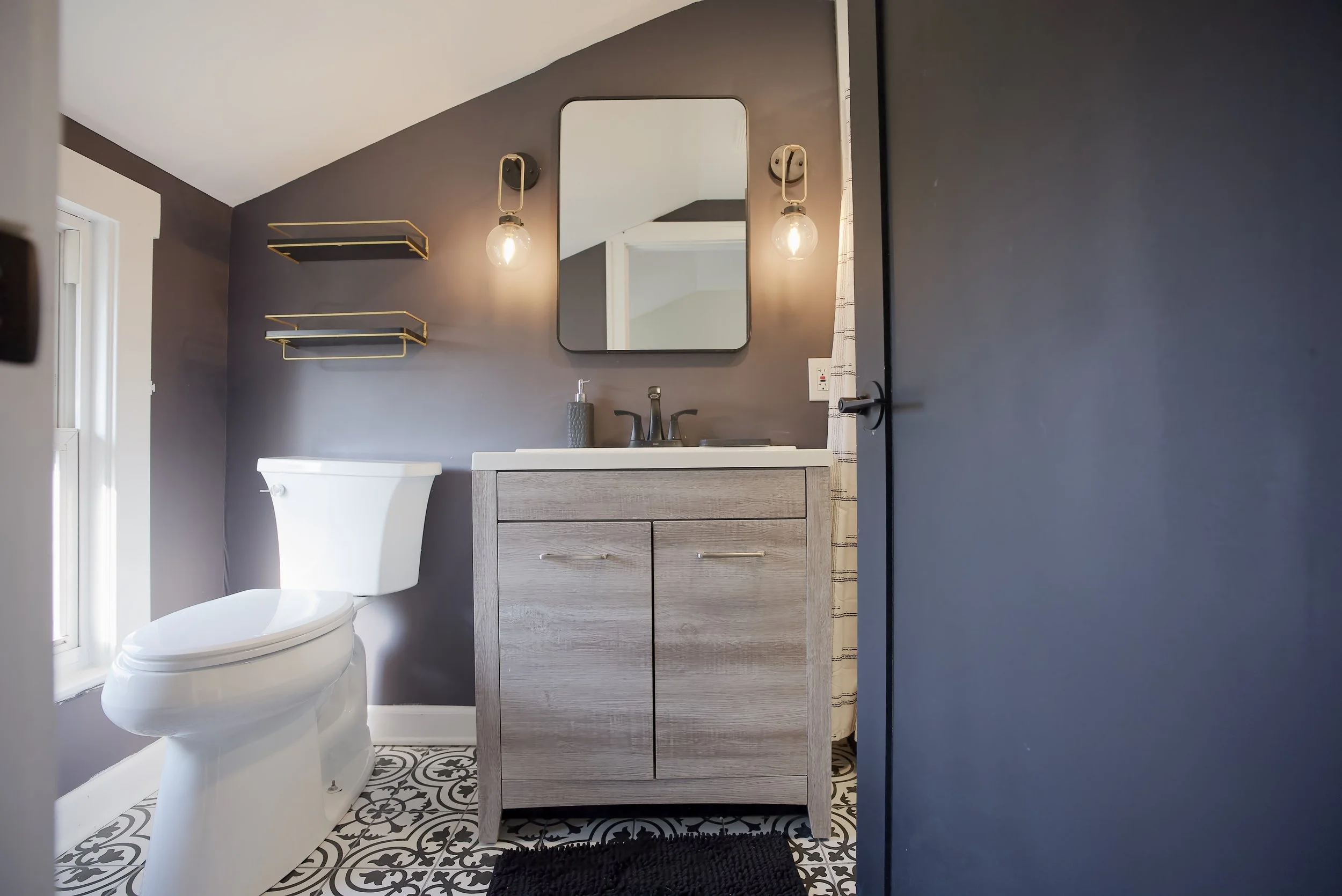 A bathroom with dark gray walls, a white toilet, a wooden vanity with a sink, a mirror above the sink, two exposed bulb light fixtures, and decorative black-and-white patterned floor tiles.
