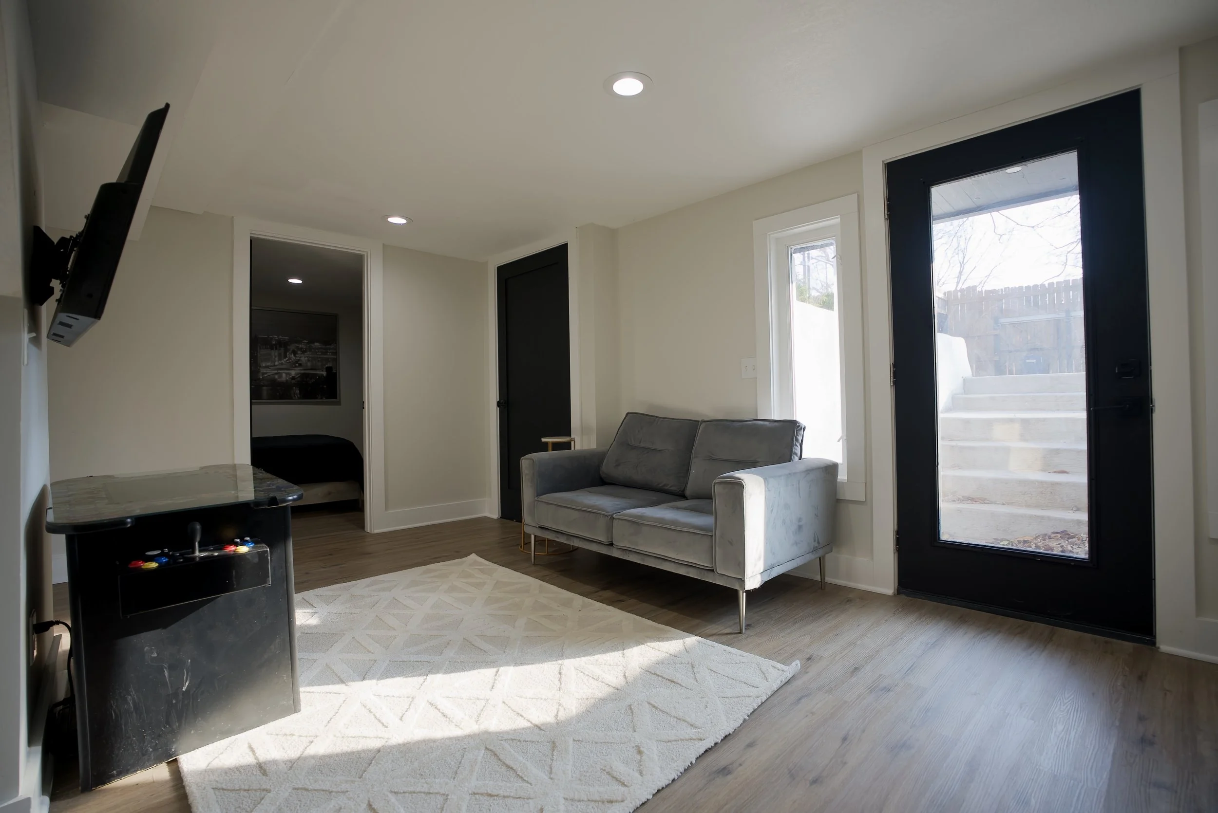 Living room with a gray sofa, a white rug, a black door, a small side table, and a large window with a door leading outside to stairs.