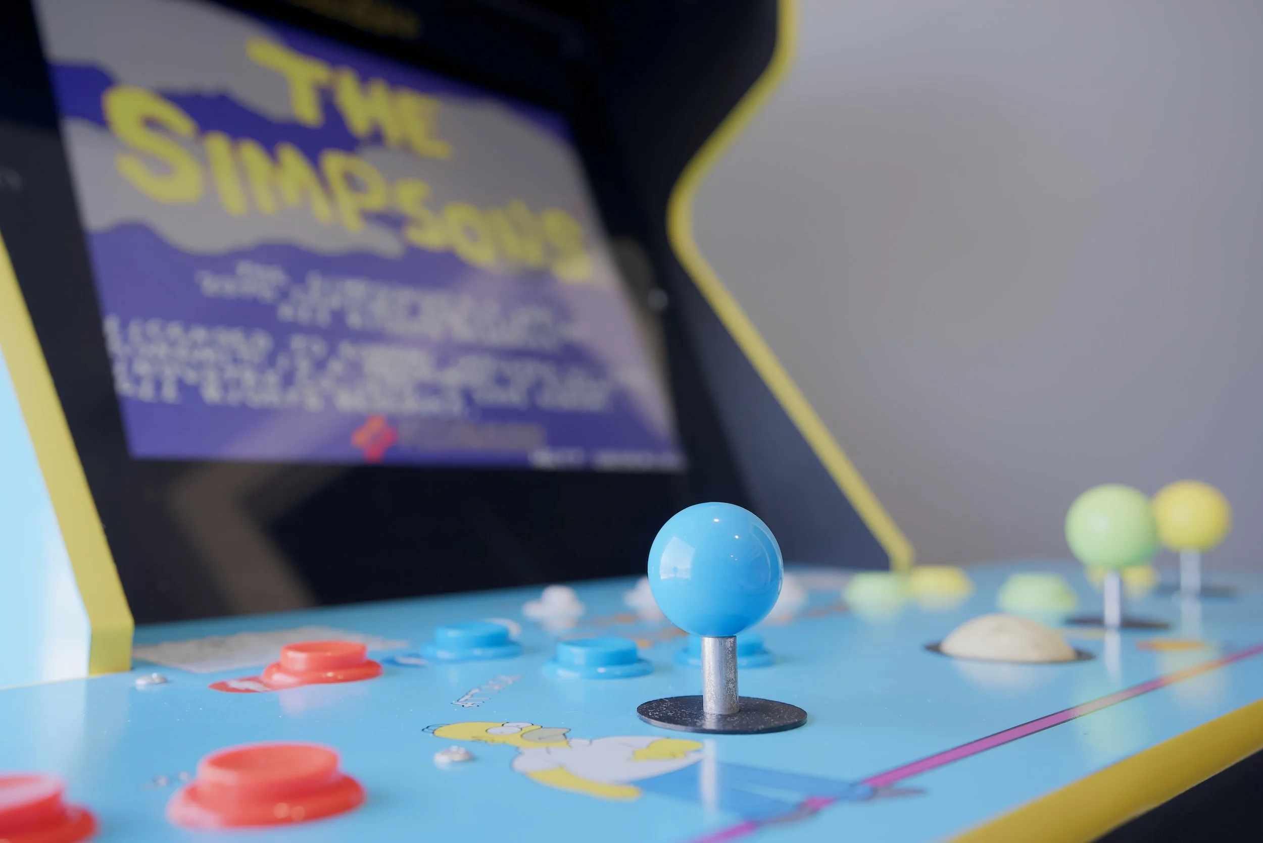 Close-up of a vintage arcade game controller with a blue joystick, red, blue, and green buttons, and a screen in the background displaying blurred text and graphics.