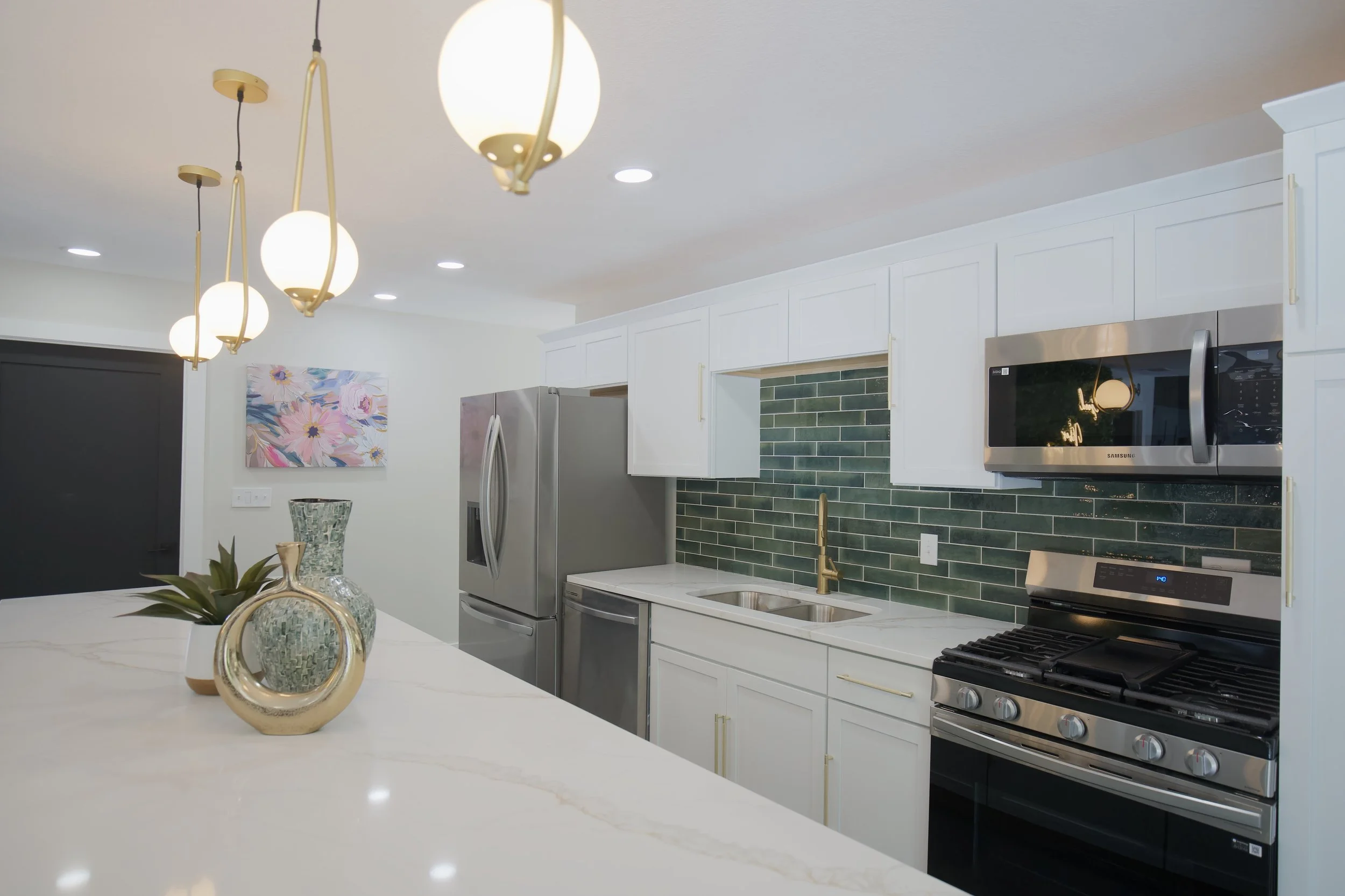 Modern kitchen with white cabinets, green tile backsplash, stainless steel appliances including a refrigerator, microwave, and oven. Features pendant lighting and decorative vases on a marble countertop. Artwork on the wall, ceiling lights, and a dar