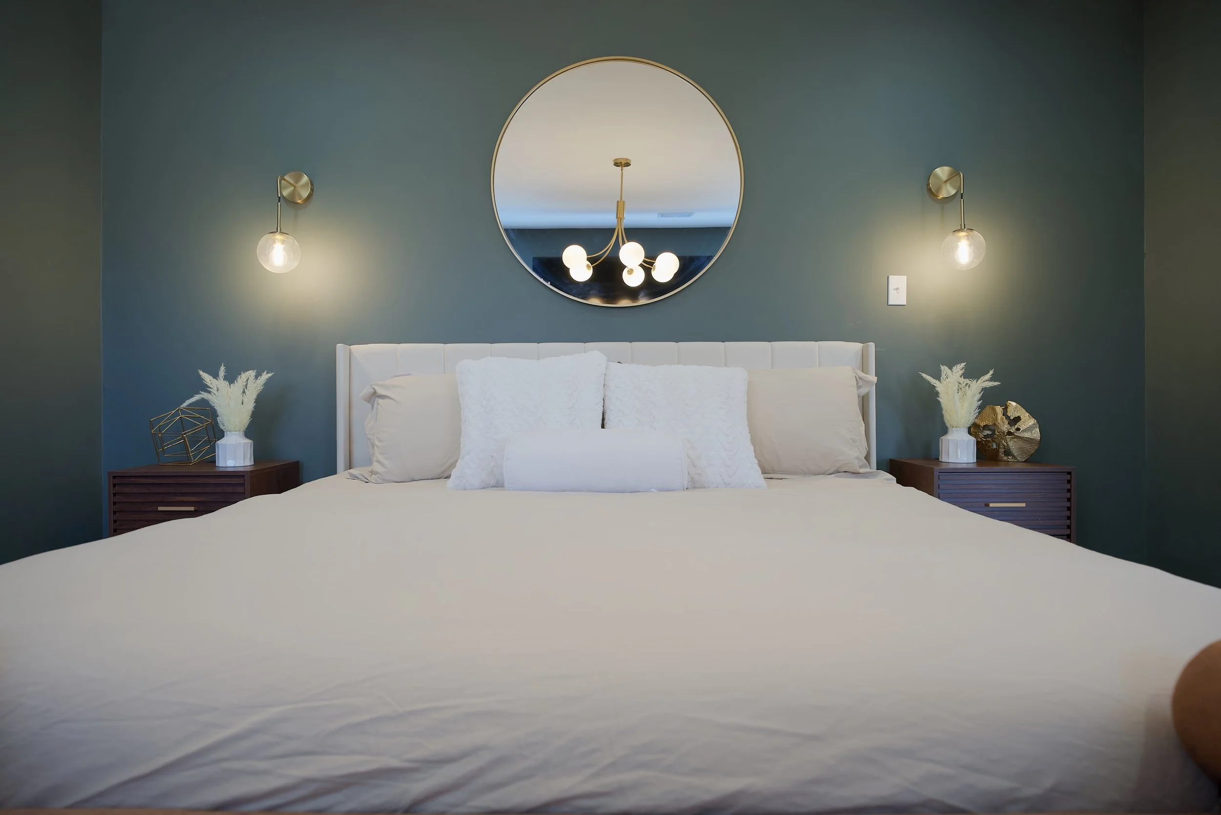 Modern bedroom with a large bed featuring white bedding and pillows, a circular mirror above the headboard, dark green wall, two matching wooden nightstands with decorative items, wall-mounted lights, and a chandelier reflected in the mirror.