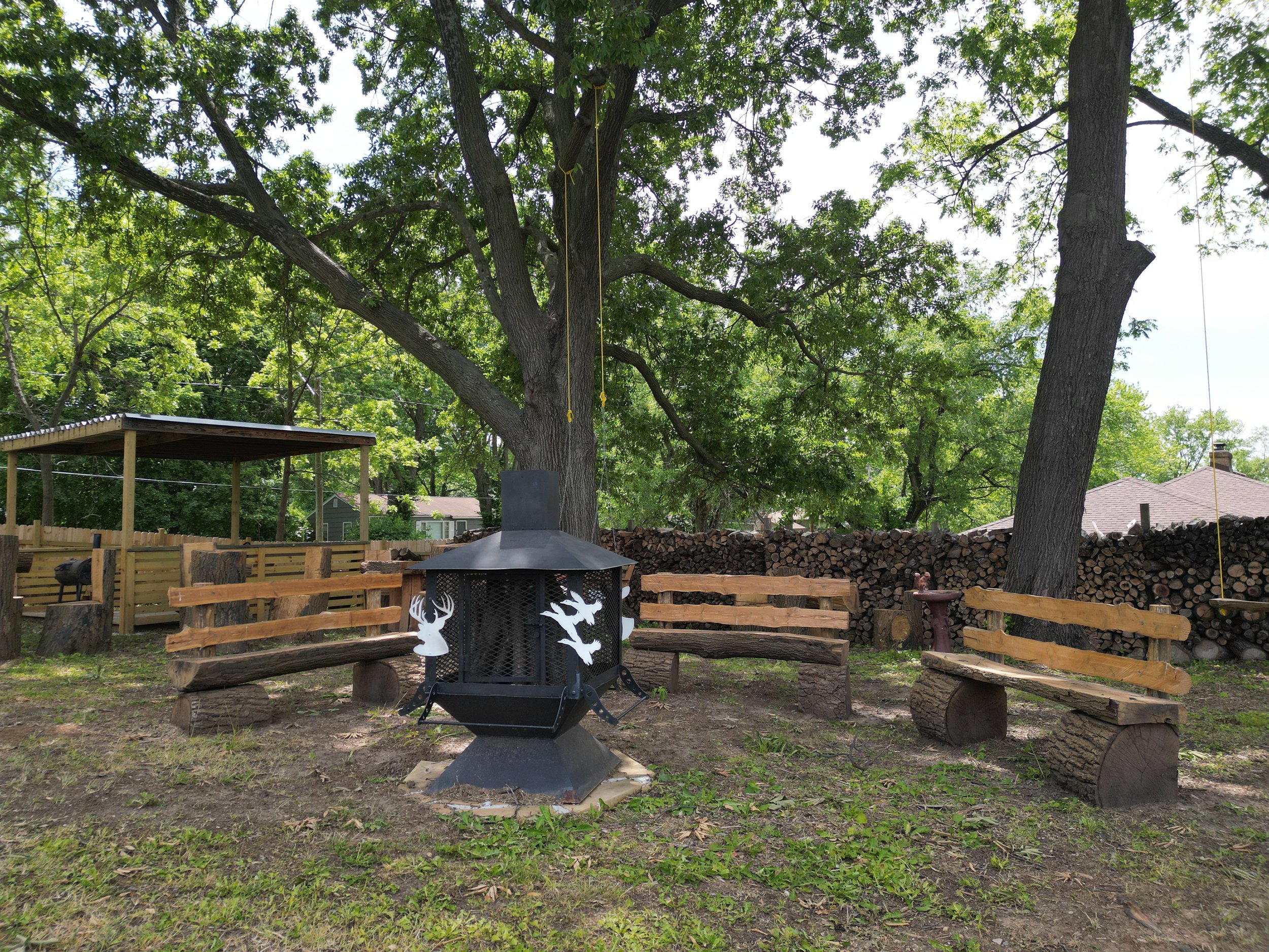 Backyard with large trees, wooden benches, and a black metal fire pit decorated with paper cutouts of deer and birds. In the background, a woodpile and a fenced area with a covered sitting space are visible.