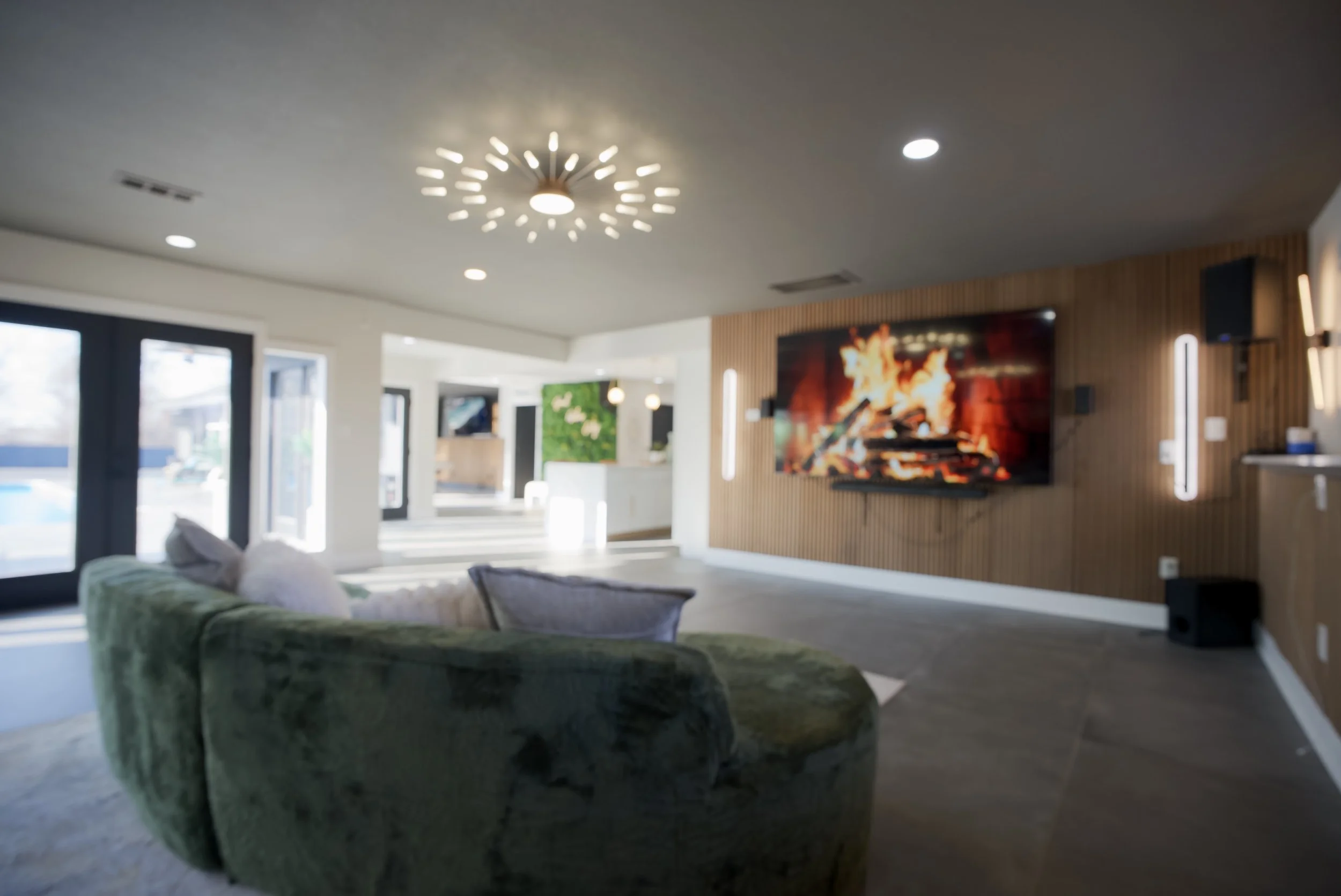 Modern living room with a green sofa, a large flat-screen TV showing a fire, wood-paneled wall, contemporary light fixtures, and large windows letting in natural light.