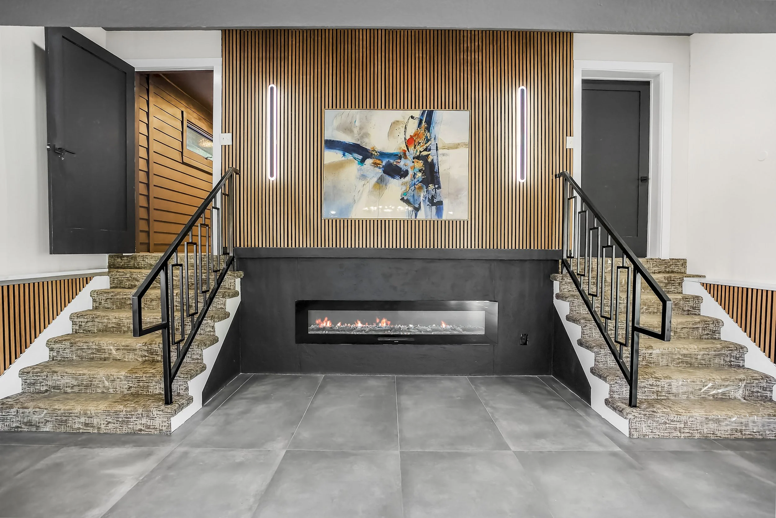 Modern interior space with a central fireplace, wooden slat wall, abstract painting, and two small staircases with black railings leading to doorways.