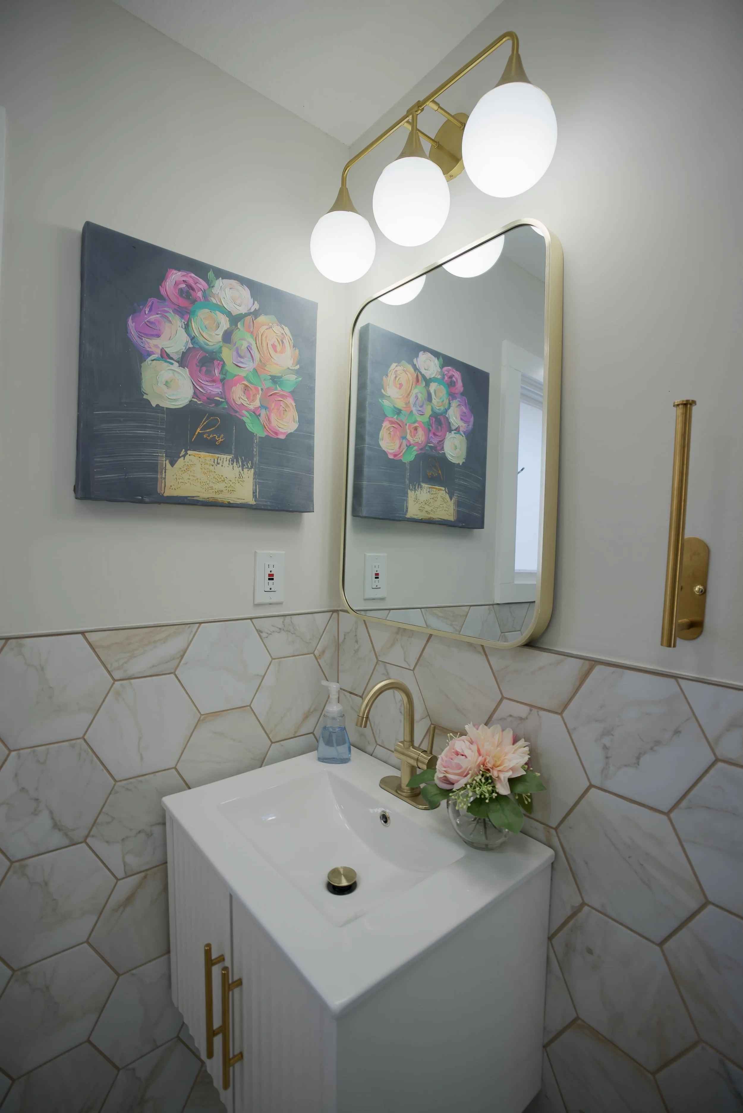 A bathroom with a white vanity, gold faucet, and a small floral arrangement. Above the vanity is a mirror, and there is a wall-mounted light fixture with four globe lights. The wall has hexagonal marble tiles on the lower half, and art prints of colo
