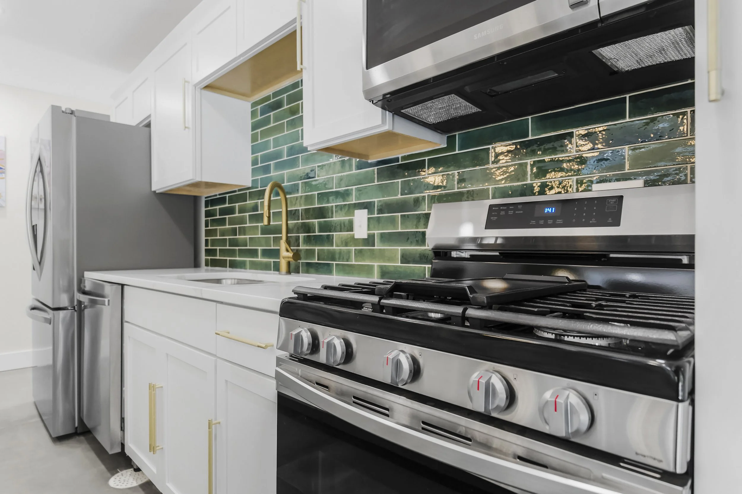 Kitchen with white cabinets, gold faucet, green tiled backsplash, stainless steel refrigerator, oven, and microwave.