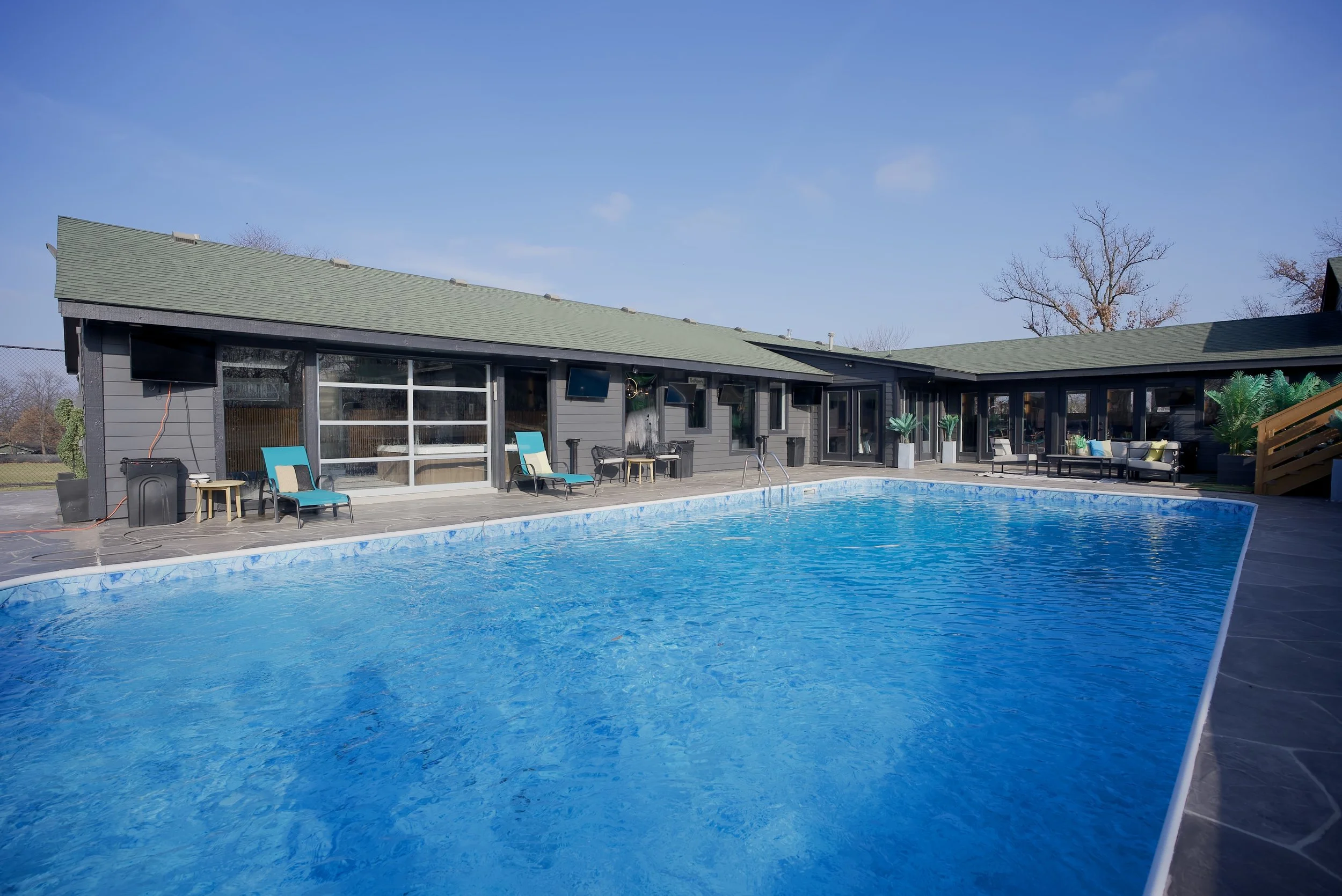 An outdoor swimming pool surrounded by a deck with chairs and tables, adjacent to a modern dark gray building with large windows, potted plants, and seating areas, under a clear blue sky.