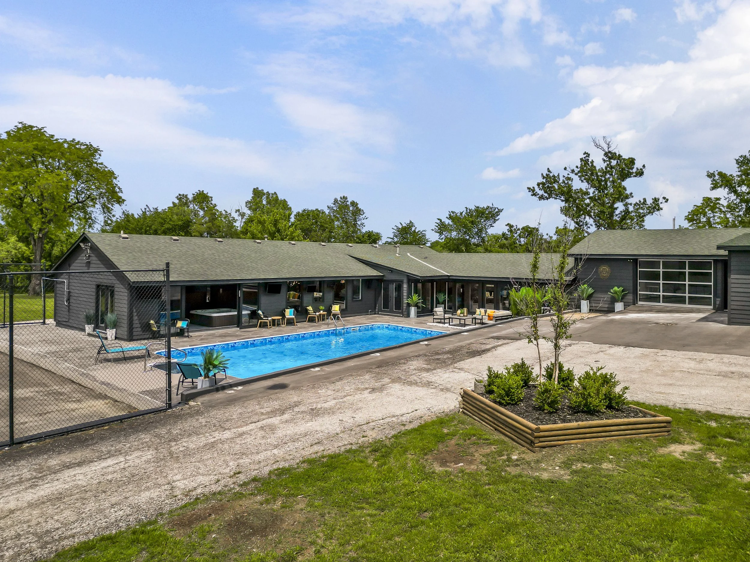Modern house with a swimming pool in the backyard, surrounded by outdoor furniture, potted plants, and a fenced area under a blue sky with clouds.