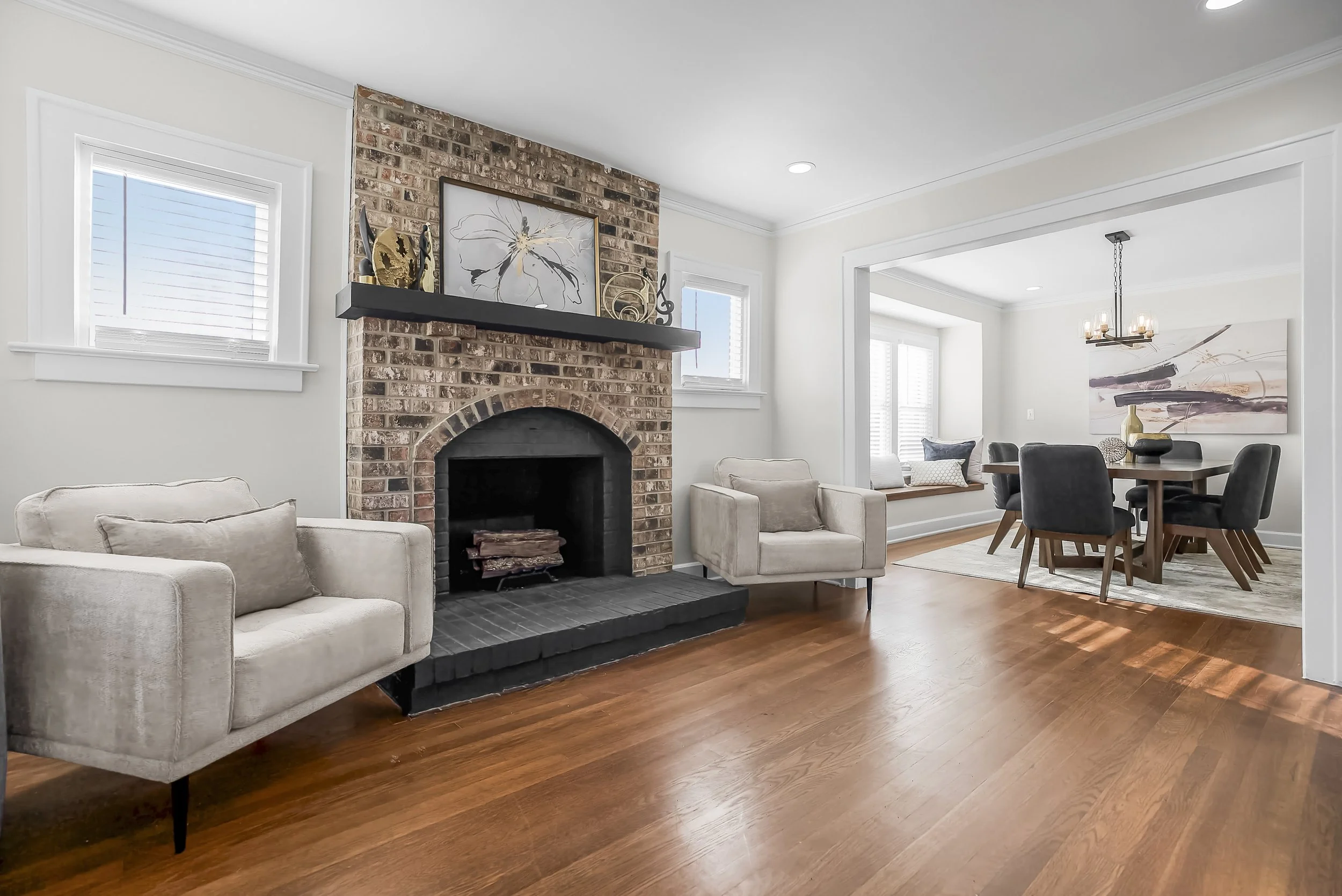 Living room with brick fireplace, beige sofas, hardwood floors, and a view into a dining area with a round table and black chairs.
